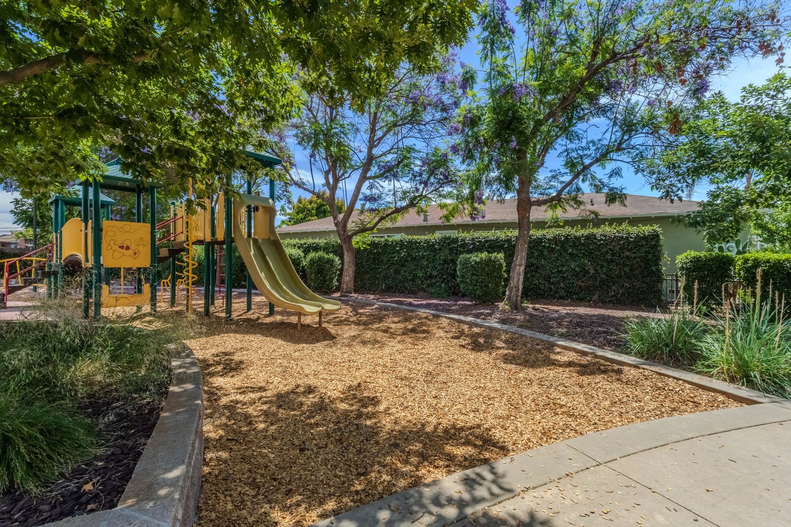 A sunny playground with slides and climbing structures surrounded by lush green trees and a mulch ground covering.