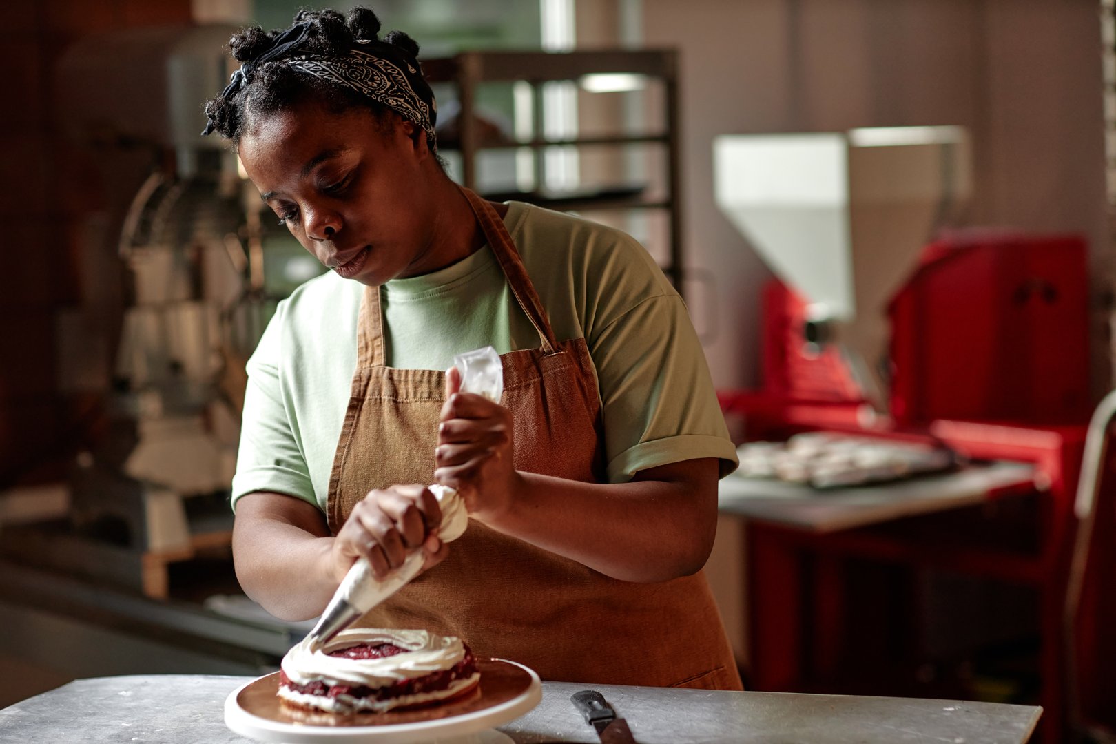 Medium shot of Black female pastry chef in apron layering cake with white buttercream using piping bag on working table in small patisserie kitchen, copy space
