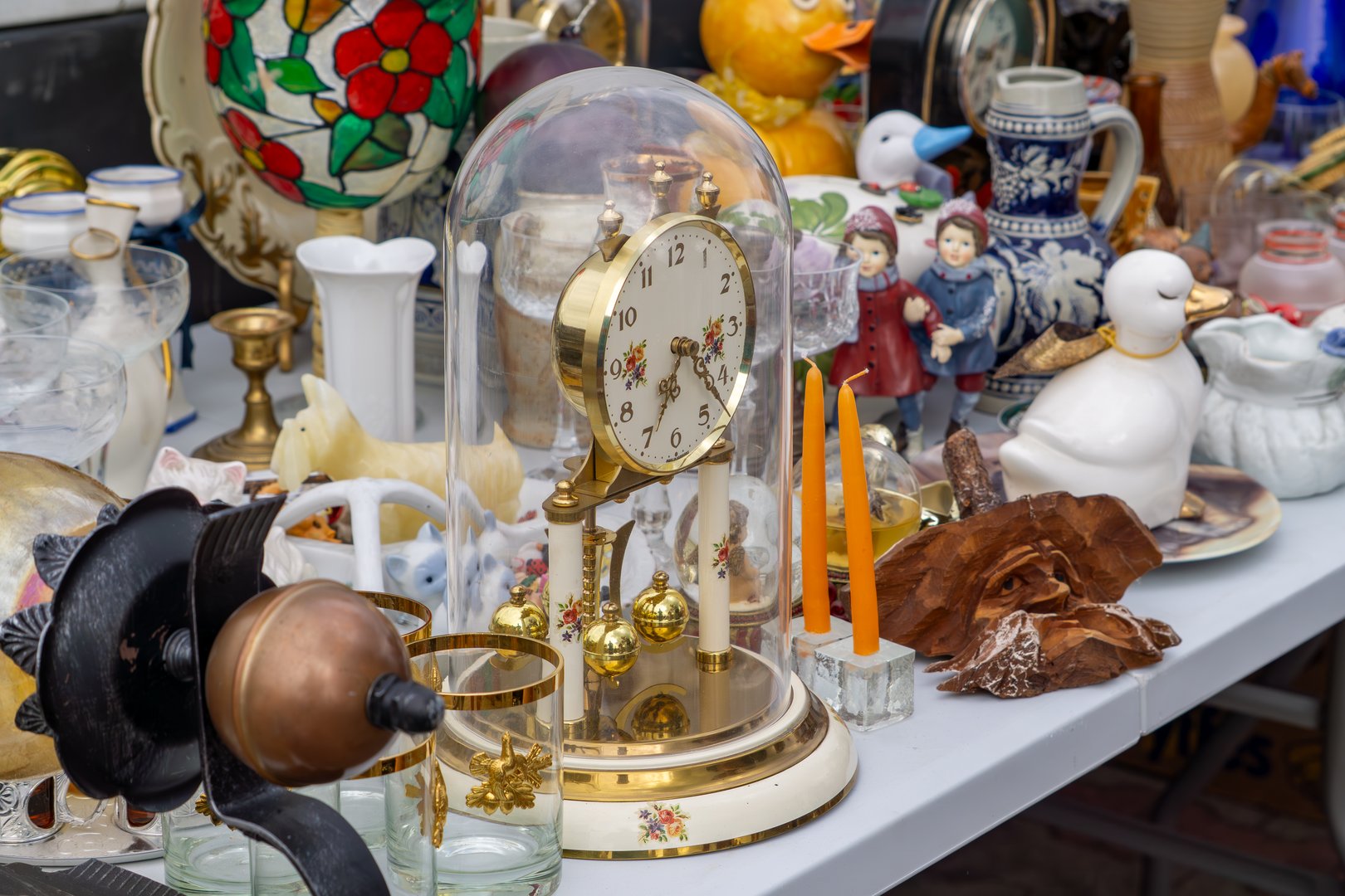 Golden mantel clock under a glass dome surrounded by figurines, glassware, and ceramics on an antique market stall. Colorful vintage assortment.