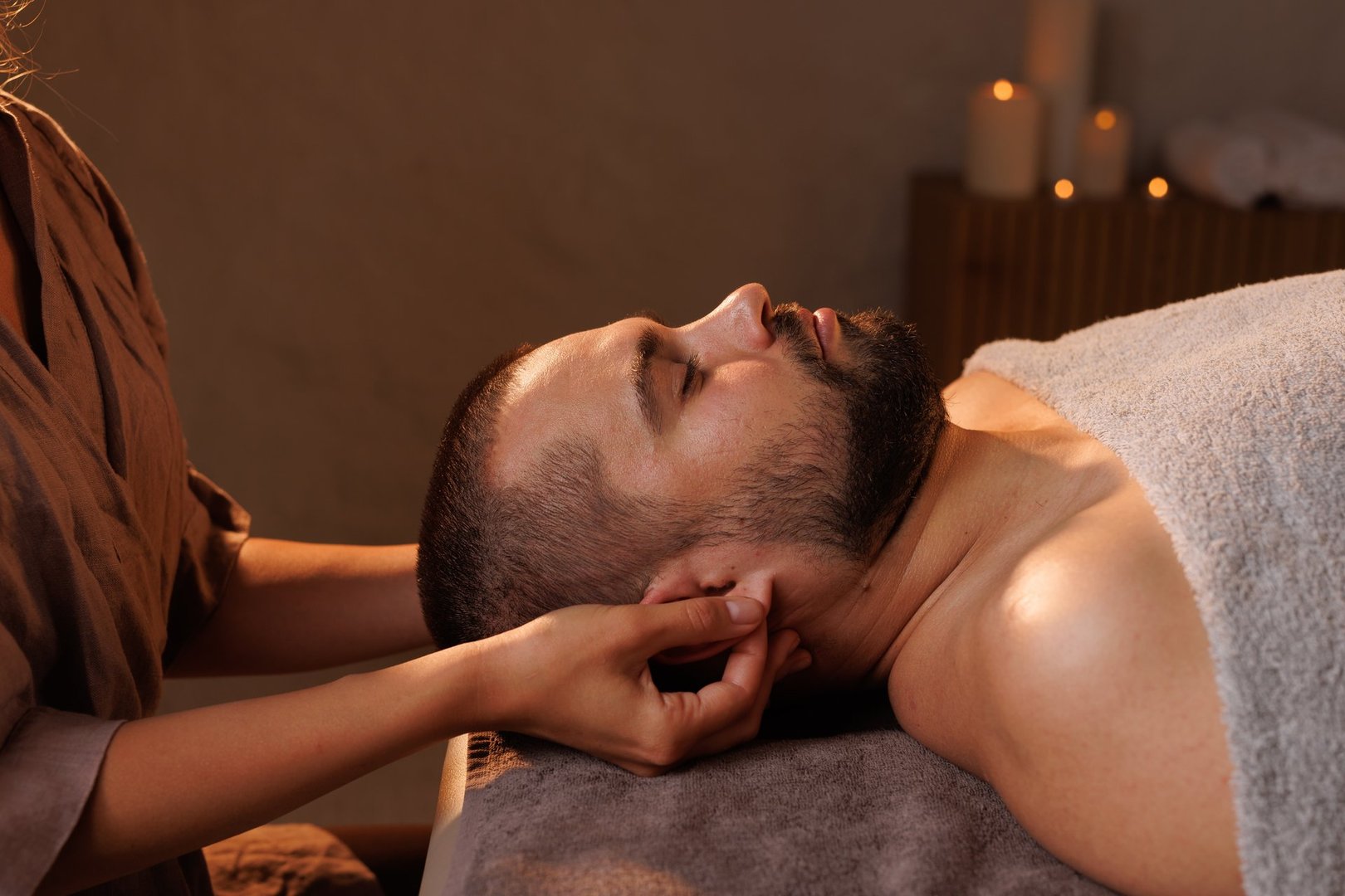 masseuse massages ear lobes of a man in a romantic warm atmosphere, relaxing massage surrounded by candles