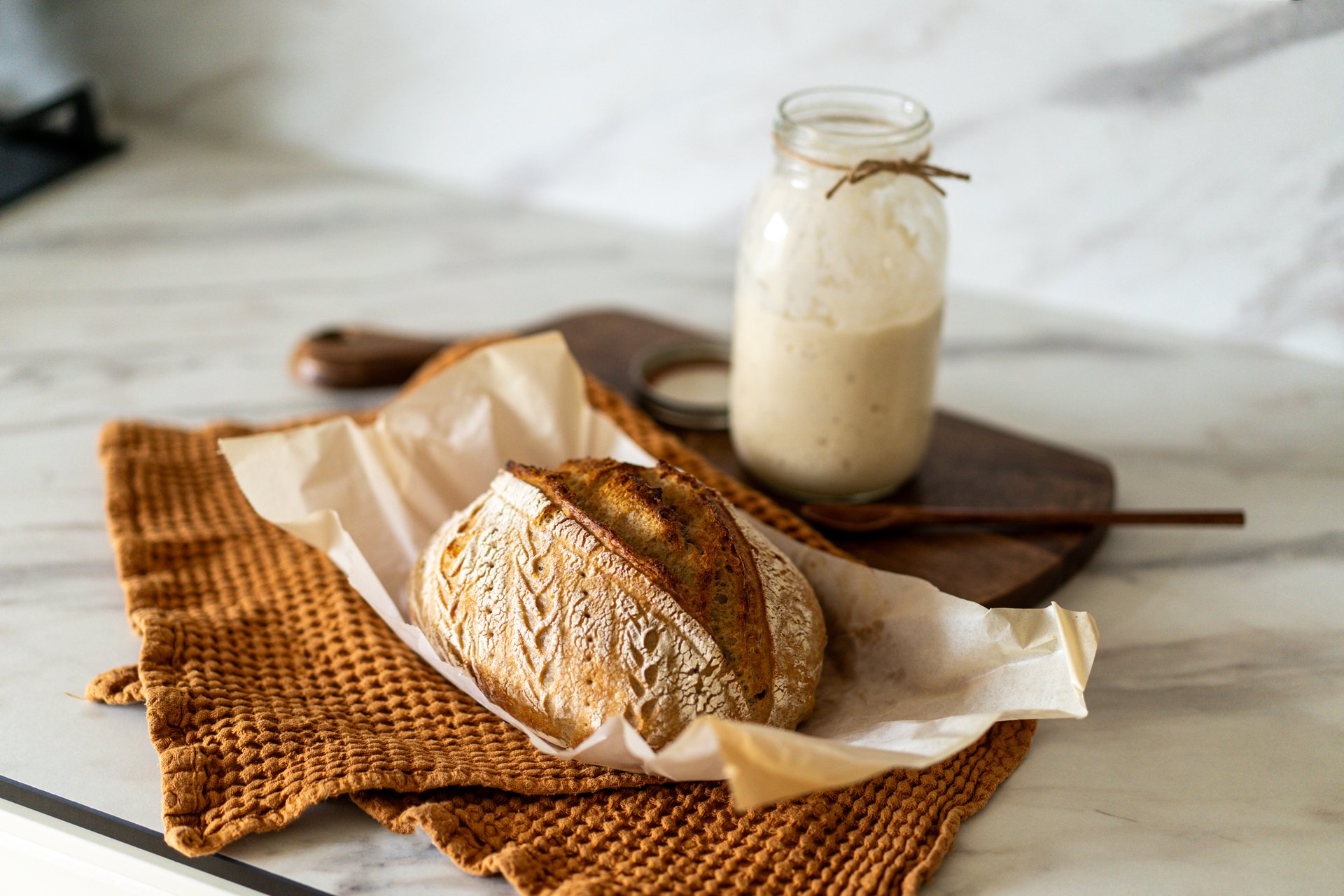 Crispy sourdough bread sits beside a glass jar filled with active starter, accompanied by a rustic cloth and a wooden board in a warm kitchen atmosphere.