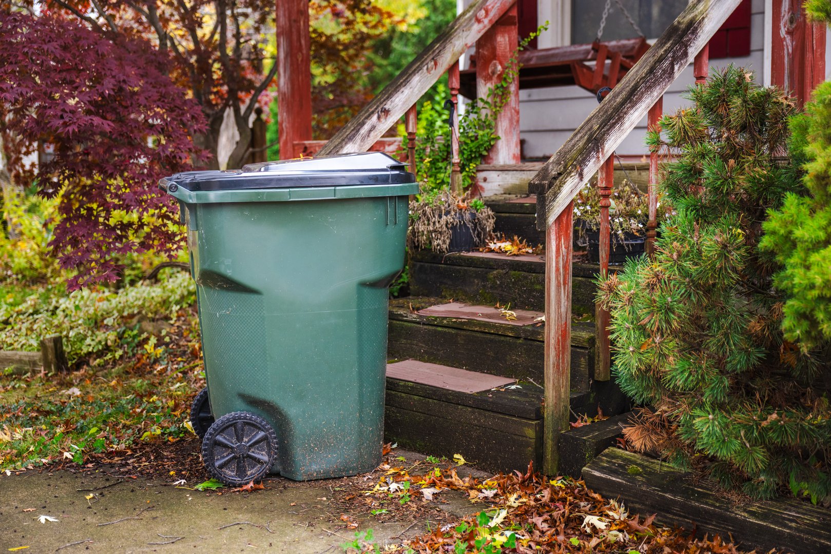 Large green garbage bin on wheels standing near the front steps of an old wooden porch surrounded by autumn foliage