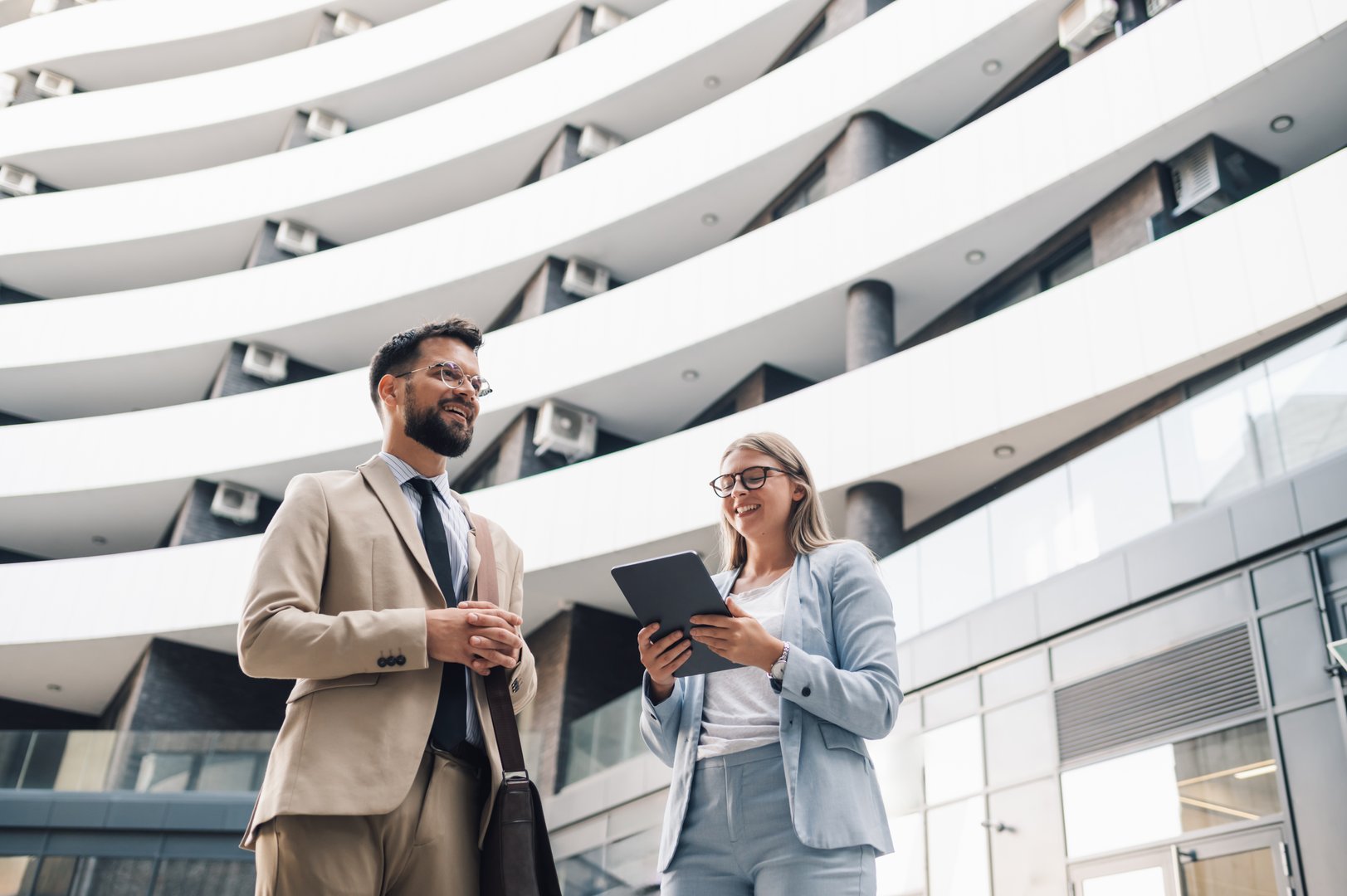 Two young professionals standing in front of a modern office building, engaging in conversation while looking at a tablet