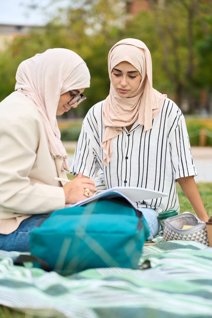 Two young adult muslim women, wearing hijabs, learning and reviewing notes while sitting on a blanket in a park