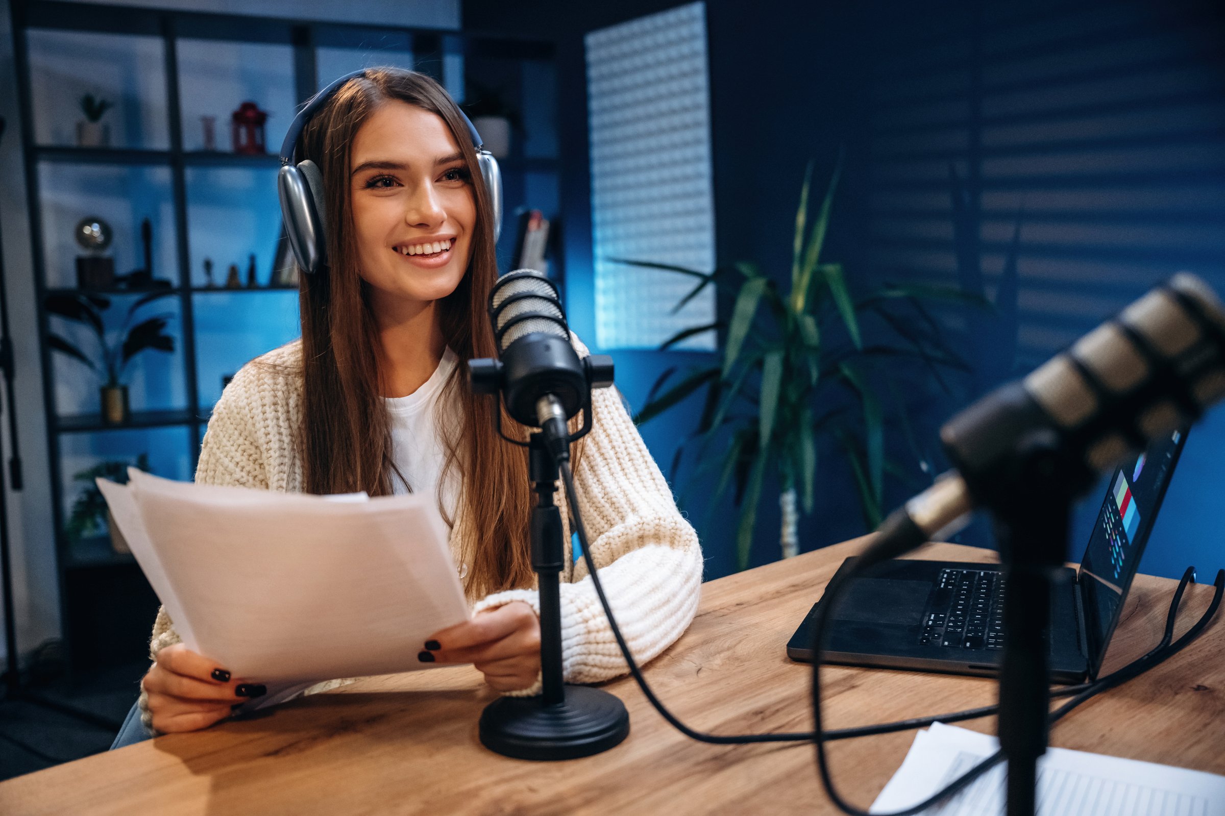 Front view, sitting. Woman is hosting radio podcast with microphone and laptop.