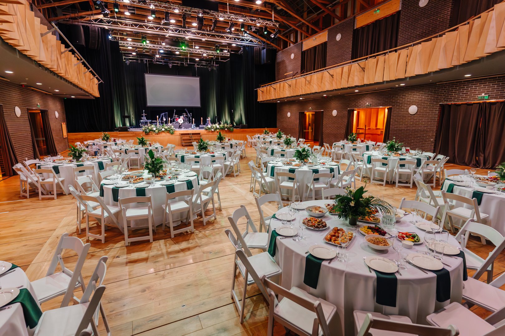 Large event hall with elegantly set round tables, floral centerpieces, food, and a stage prepared for a performance or formal gathering.