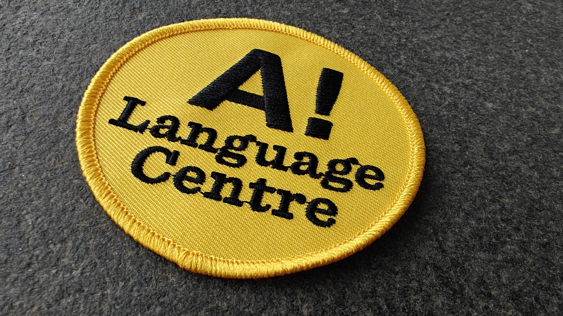 Yellow circular patch with A! Language Centre embroidered in black text on a dark gray background.