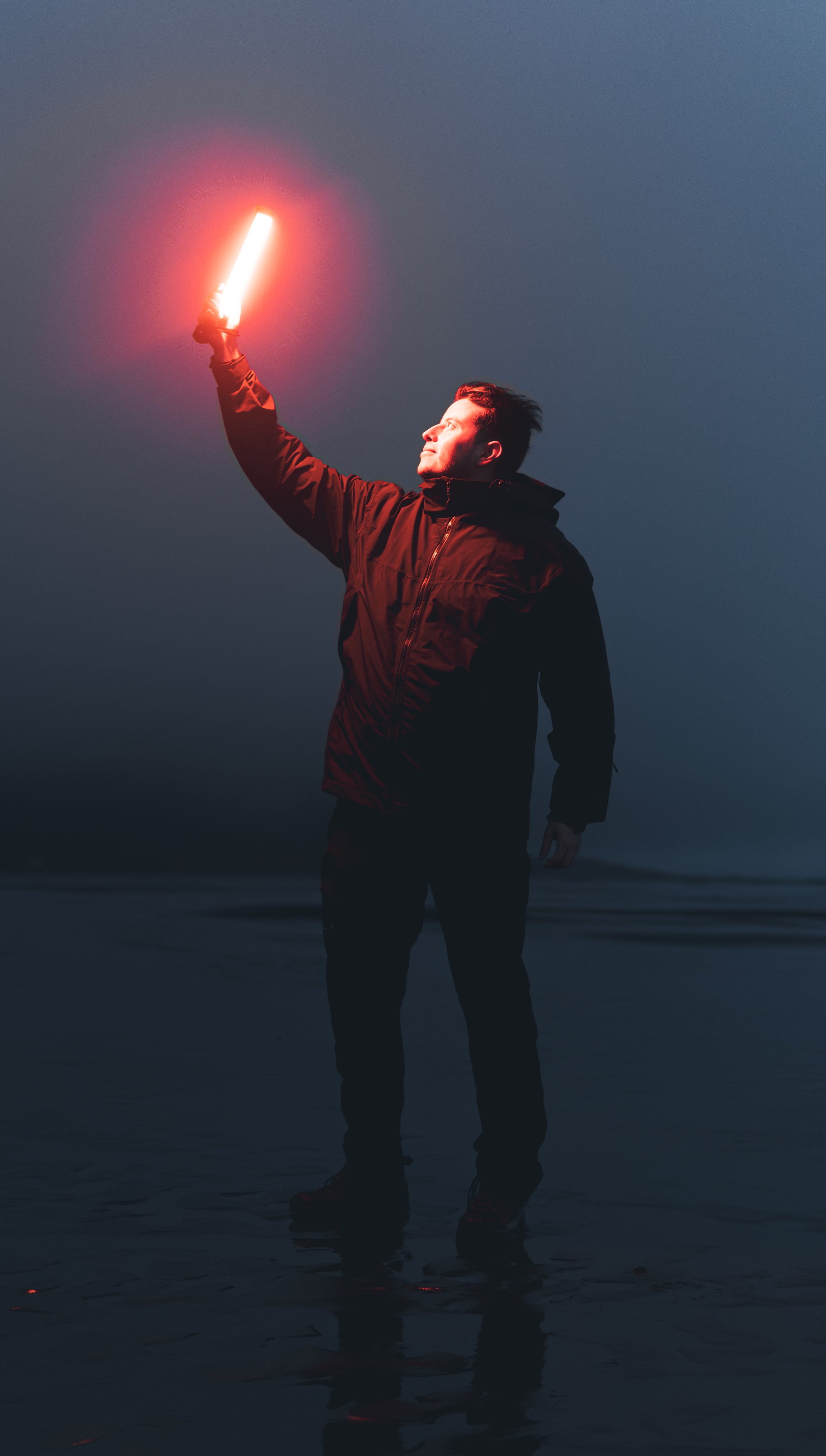 Man stands alone on a wet beach at night, holding a red flare, gazing up with hope in his eyes, surrounded by darkness and danger
