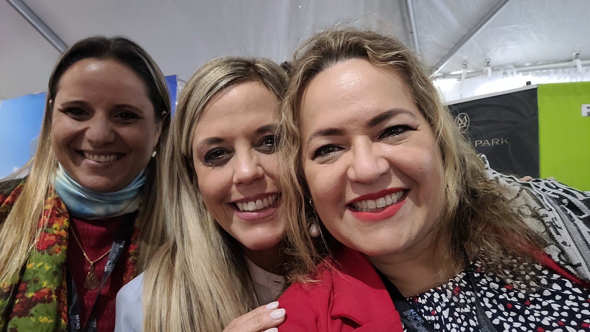 Three women smiling at the camera inside a tent, wearing lanyards, with colorful clothing and a green background.