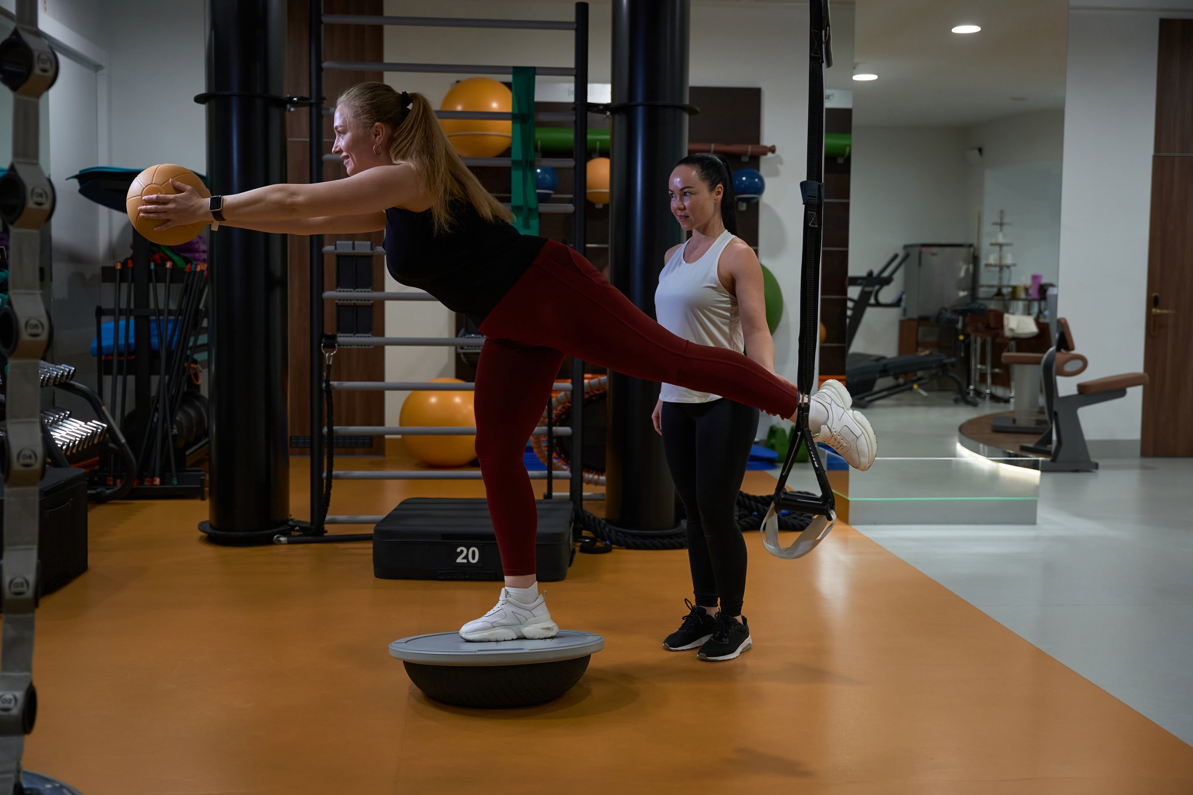 Woman performs balance exercises with a ball on a special platform, she is under the supervision of a fitness trainer