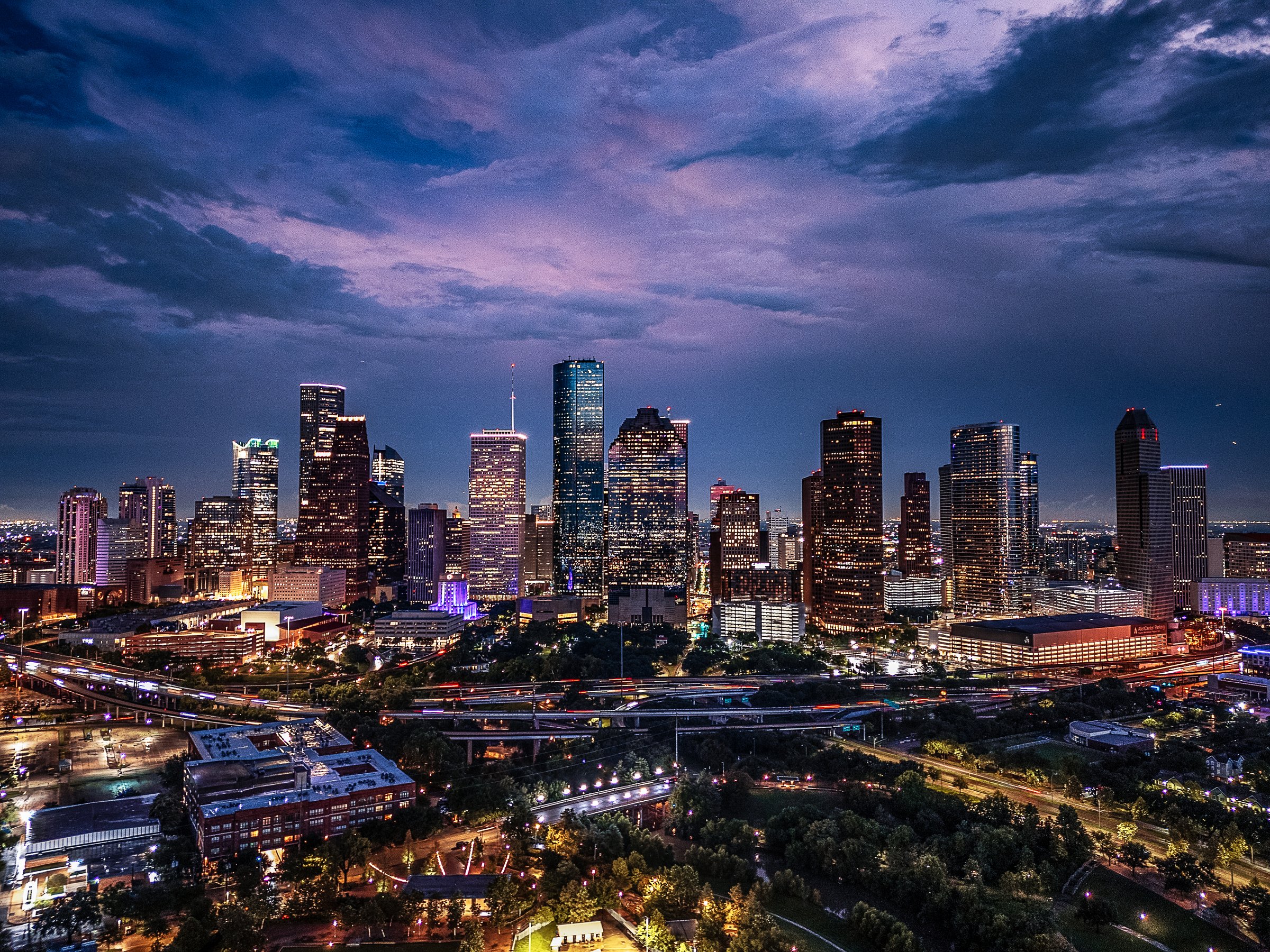 Drone aerial image of downtown Houston, Texas captured at twilight. The photograph highlights the illuminated skyscrapers, highways, and cityscape under a dramatic evening sky. A vibrant view of one of the largest cities in the United States and a major economic hub.