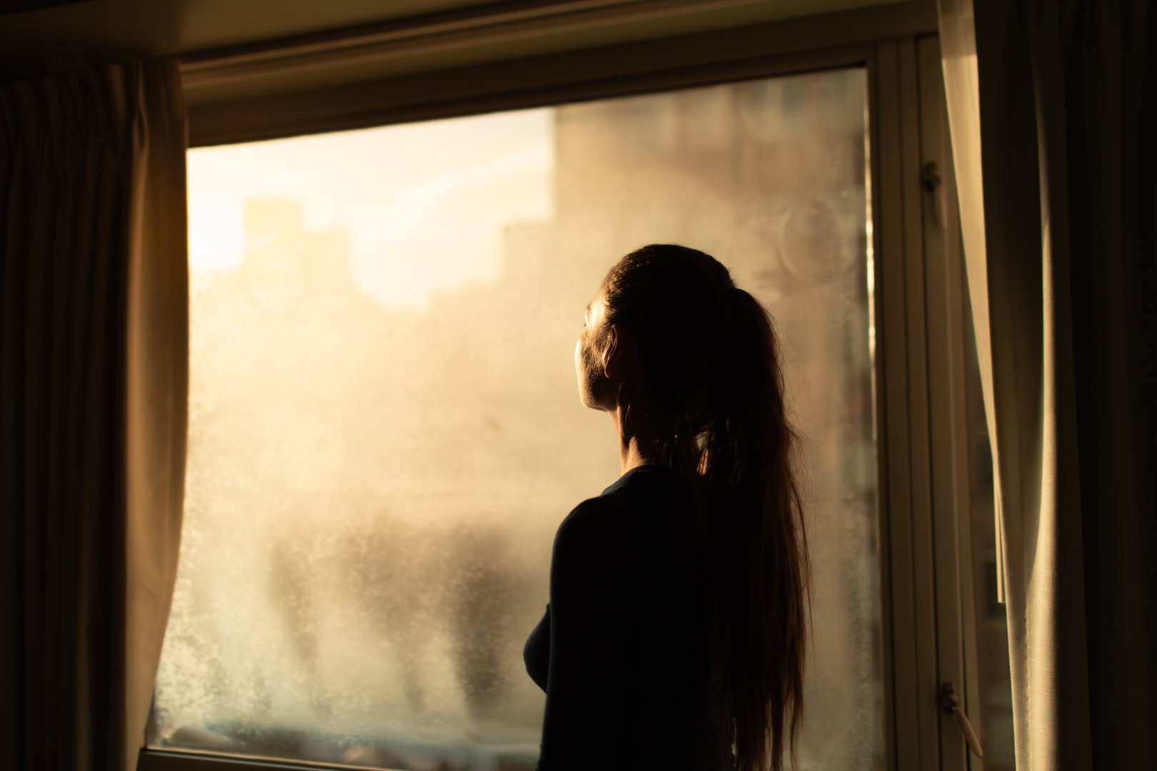 Woman in city apartment looking out her window at the light of the sun from the darkness, sadness , hope, overcoming, salvation, happiness concept.