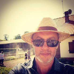 Man wearing a straw hat and sunglasses standing outside in sunny weather near a fenced area with people and a building.