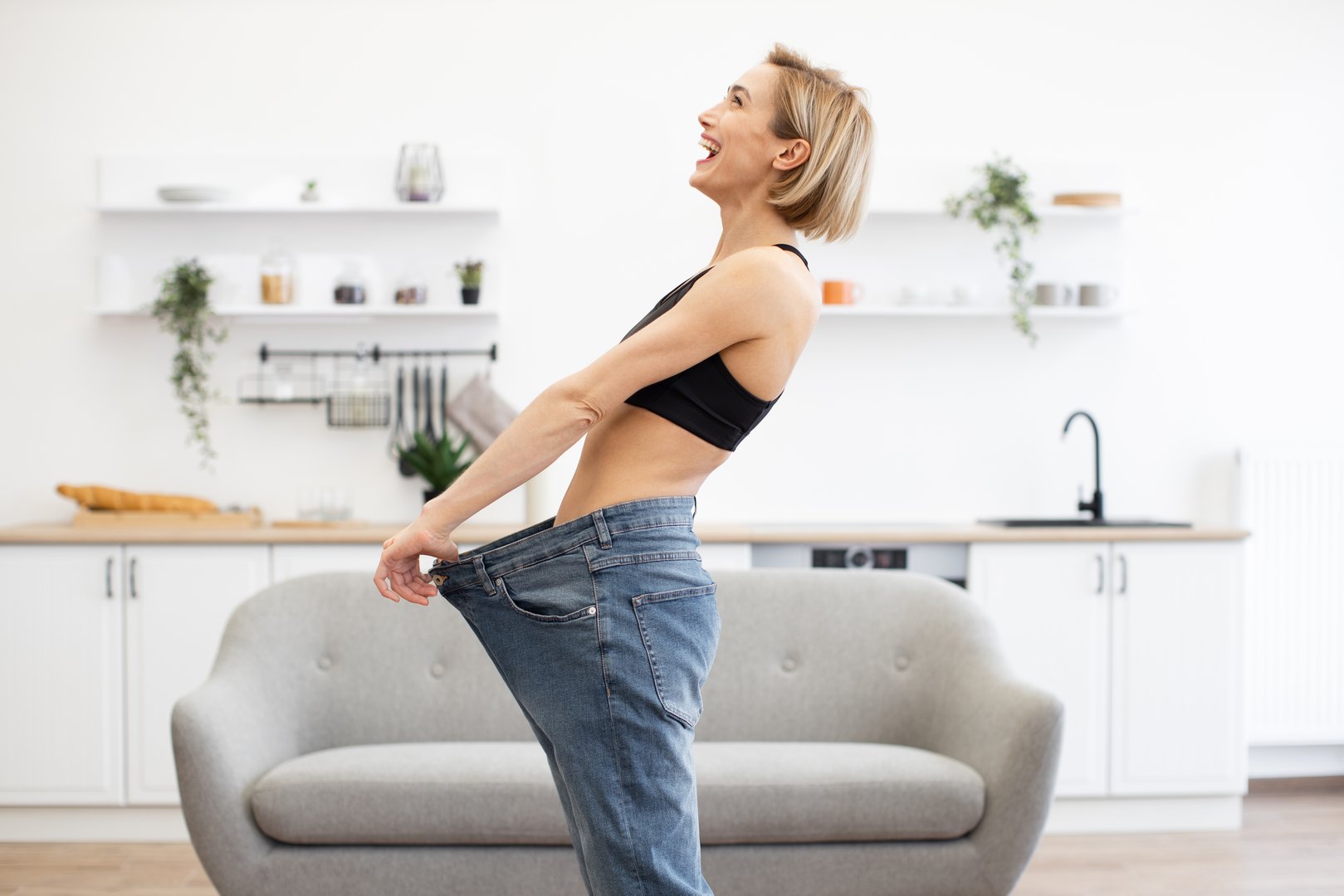 Happy woman in kitchen showing significant weight loss wearing oversized jeans. Success in dieting and fitness. Healthy lifestyle achievement.