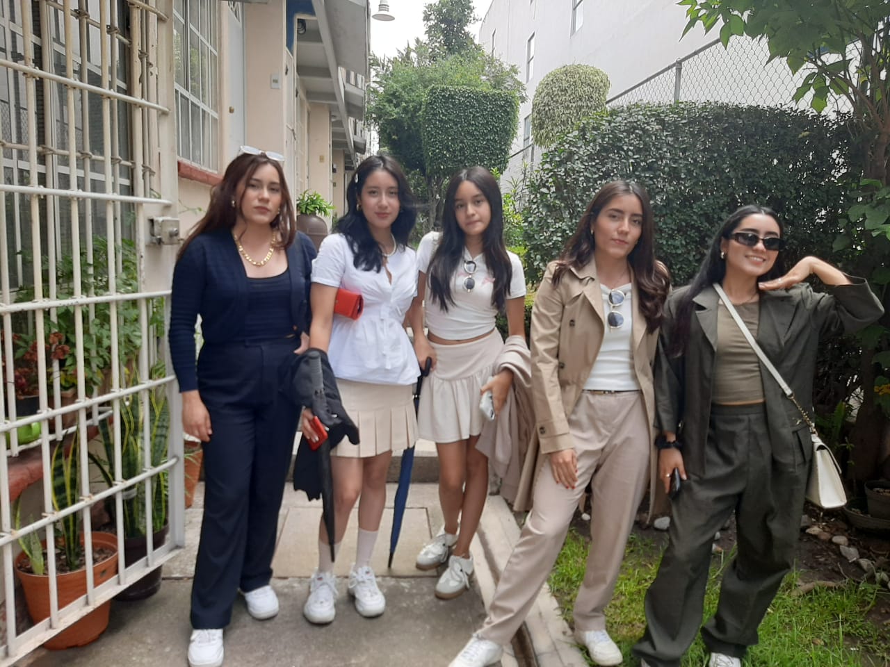 Five young women posing outdoors, dressed casually in neutral colors, standing on a pathway with greenery in the background.