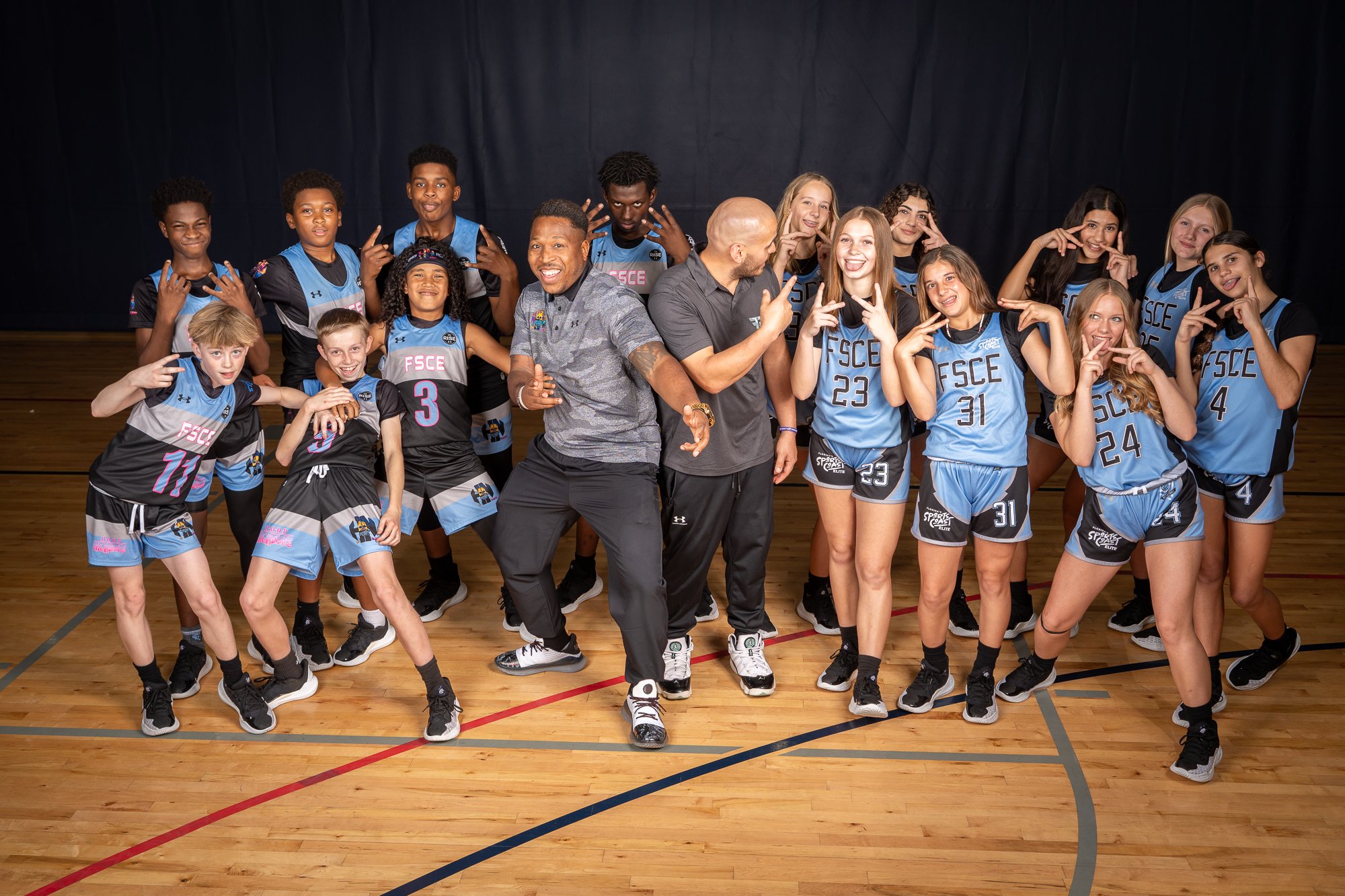 Basketball team posing with two coaches on a court, all making playful gestures, wearing blue and black uniforms.