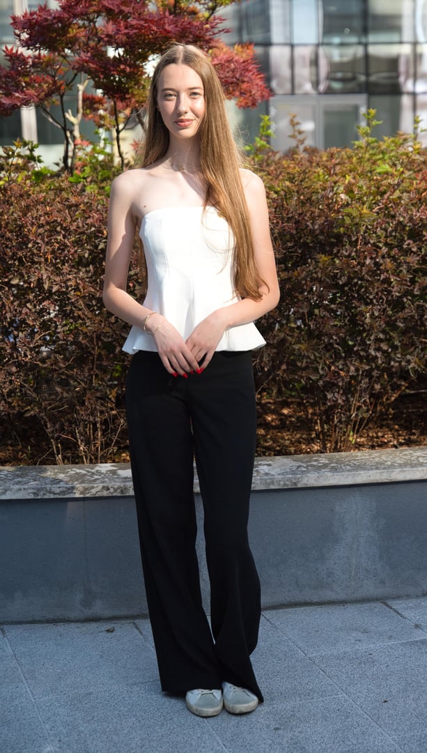 A woman with long hair wearing a black dress and smart watch stands in front of a stone wall, smiling slightly.