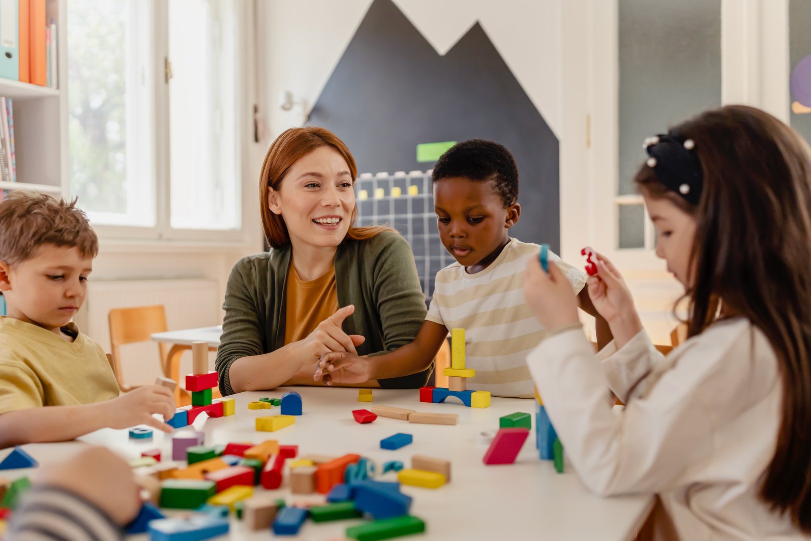 Diverse preschool children play with colorful building blocks at a classroom table, guided by a smiling teacher in a creative, inclusive early learning environment.