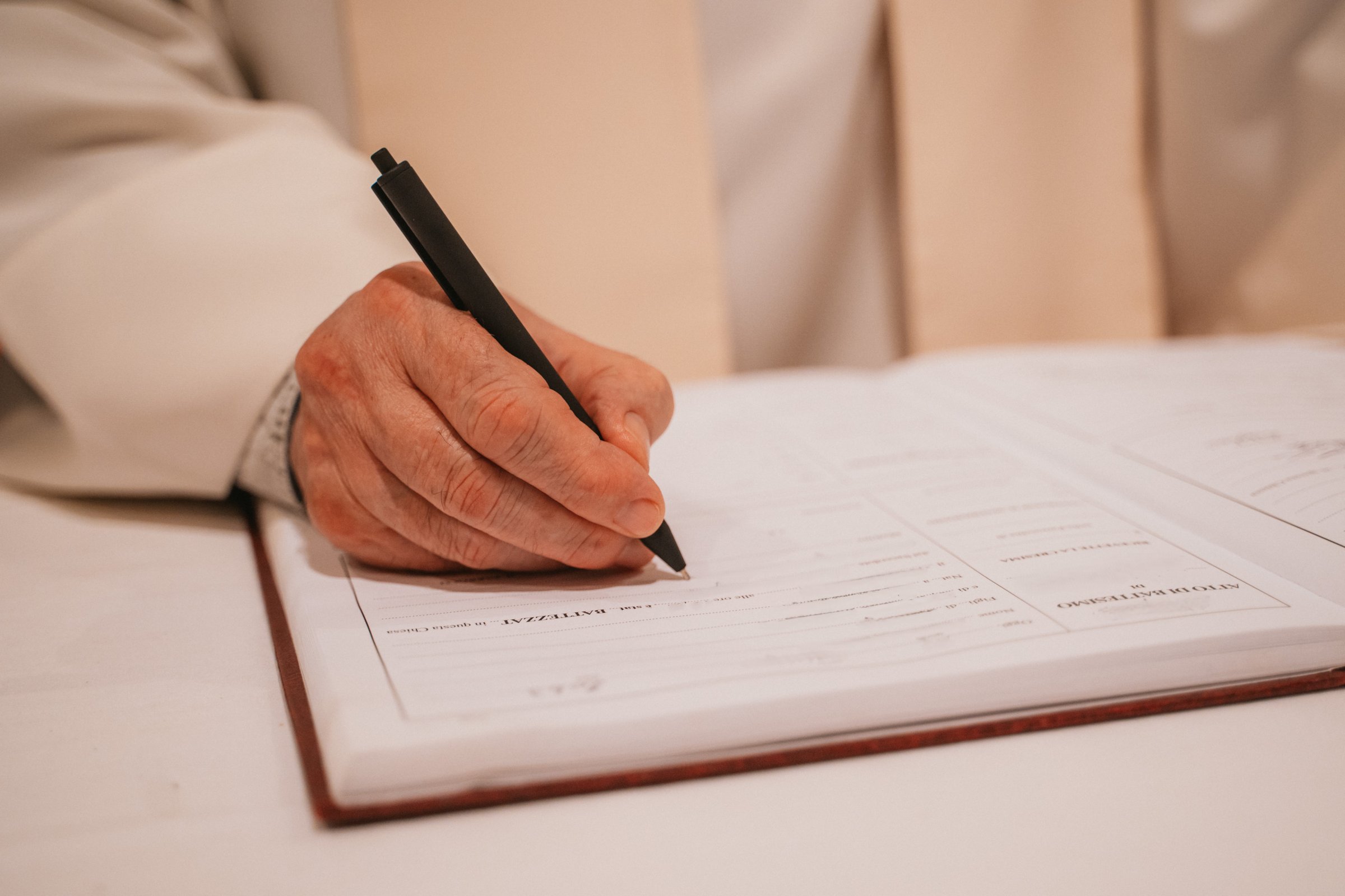 Close-up of a priest's hand writing baptism details in a church register using a pen. The page contains sections related to baptism documentation, reflecting a ritual process.