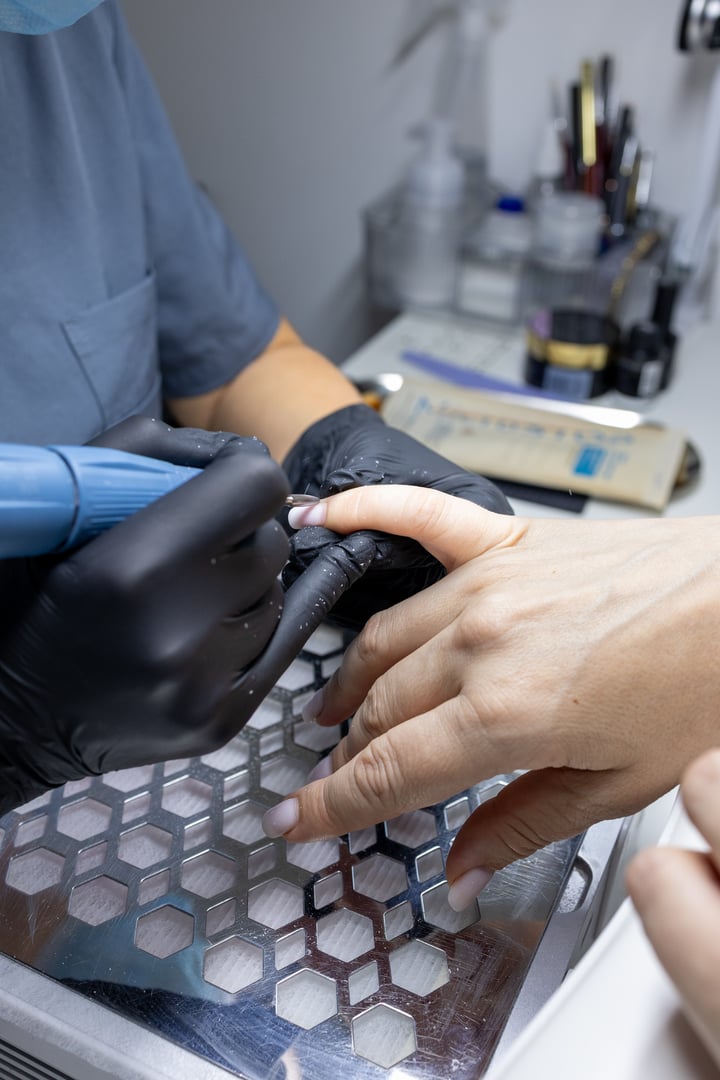 Nail technician wearing gloves performing manicure procedure with electric nail drill. Female client receiving nail care treatment in beauty salon. Professional nail service concept.