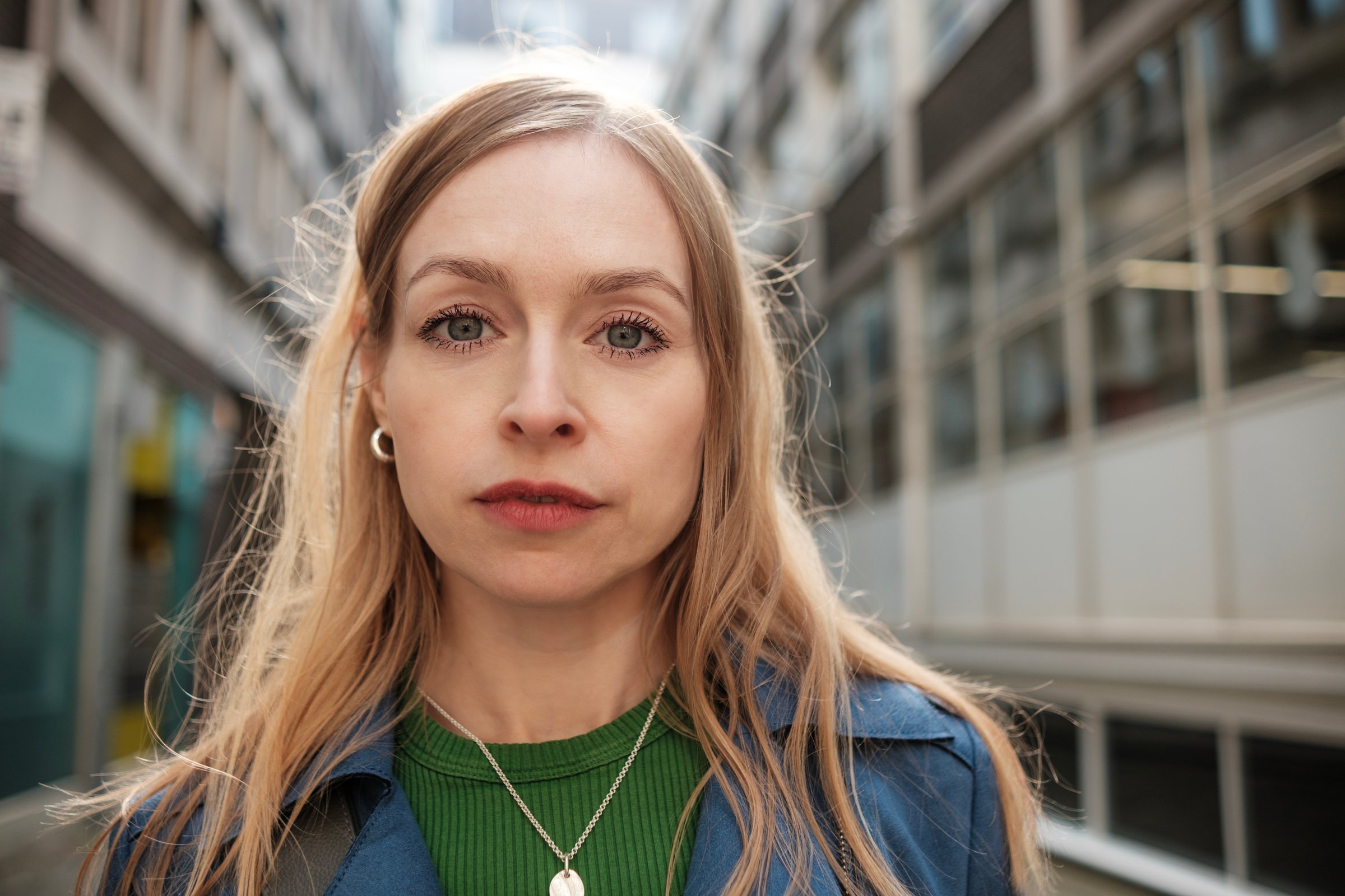 Serious young blonde woman gazing directly at the camera while standing in a bustling city street, dressed in a stylish blue jacket and a vibrant green shirt