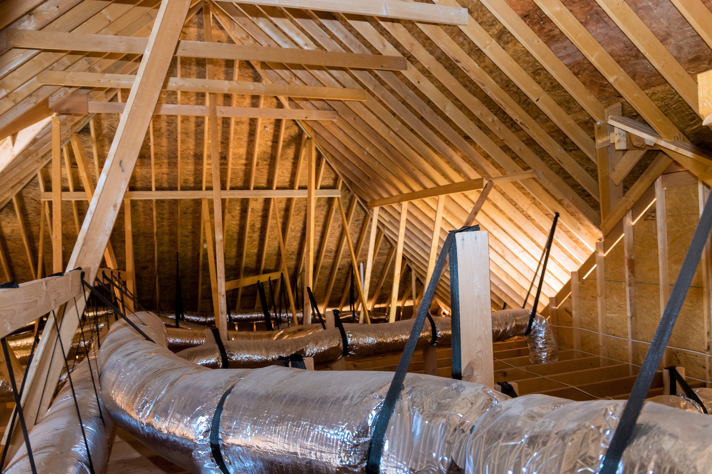 Ductwork wooden structure are visible in spacious attic, with sunlight shining through rafters.