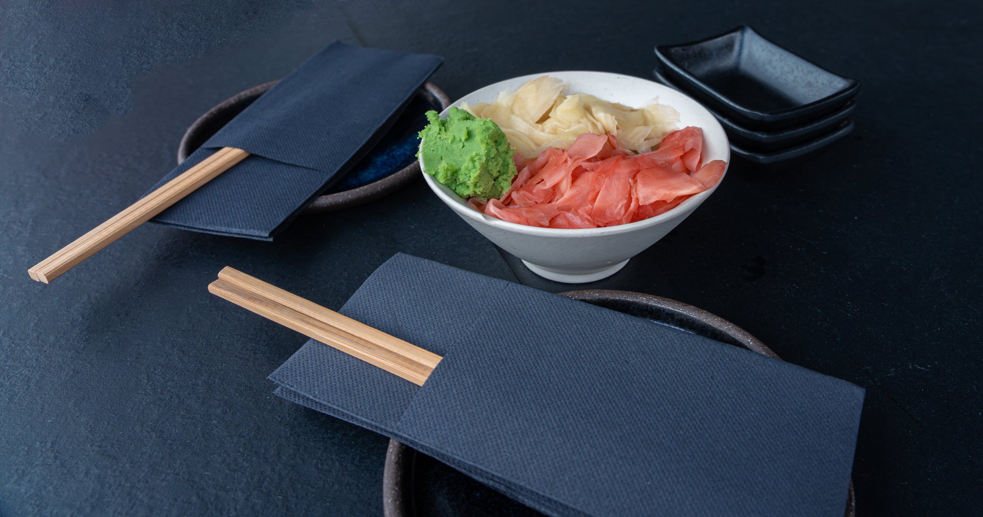 Pickled pink and white ginger and wasabi bowl on dark table, traditional Japanese condiments served with sushi, stacked sauce dishes and napkins on black background