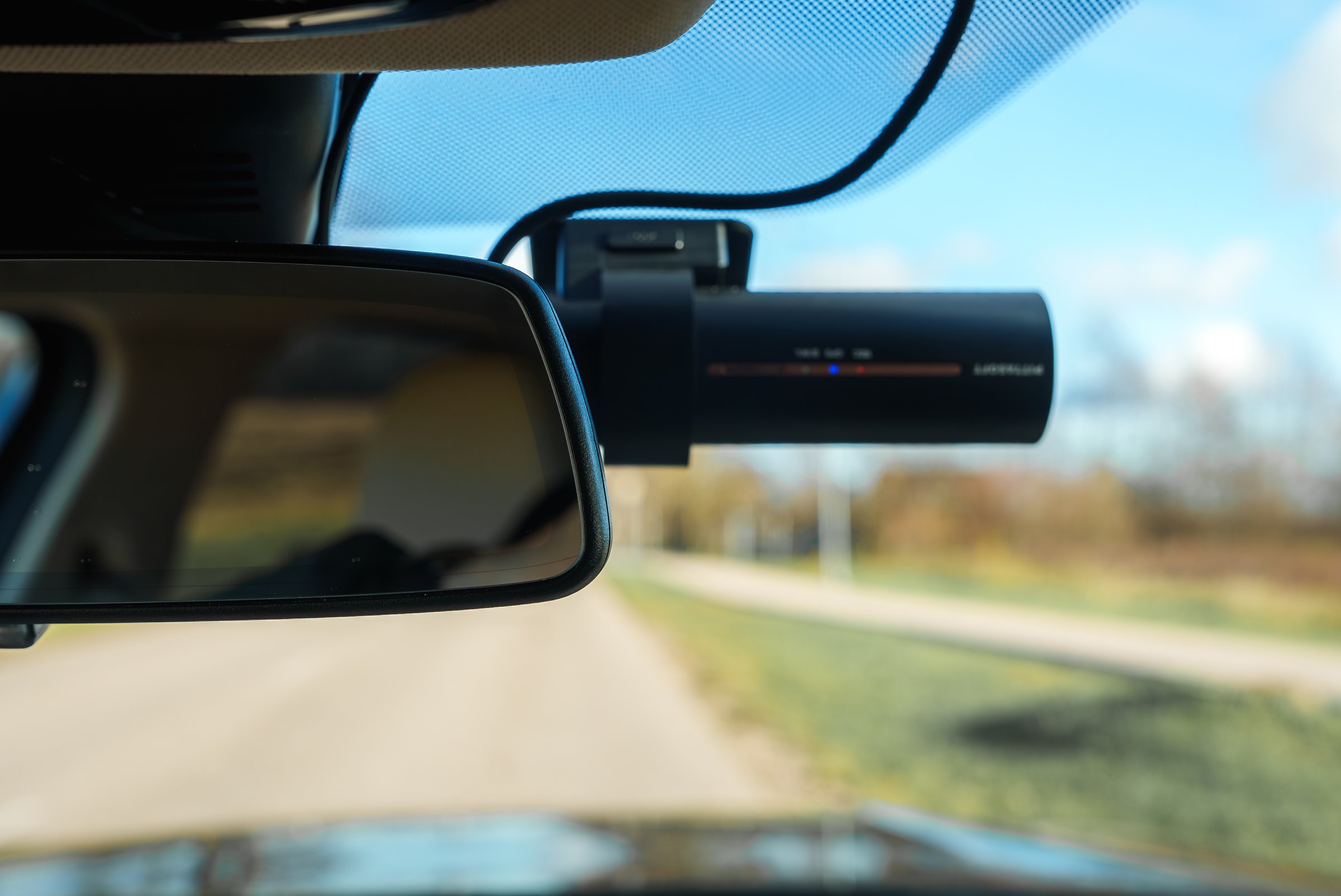 The image shows a car's interior focusing on a BlackVue dashcam and rearview mirror. Through the windshield, a rural road and trees are visible.