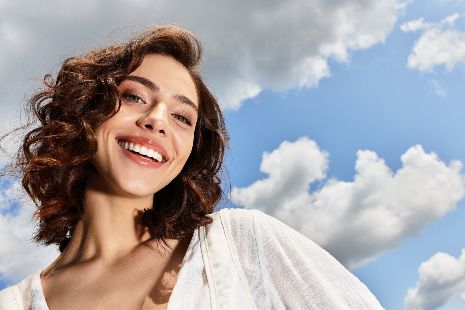A young woman with wavy brown hair smiles outdoors on a sunny day with a blue sky.