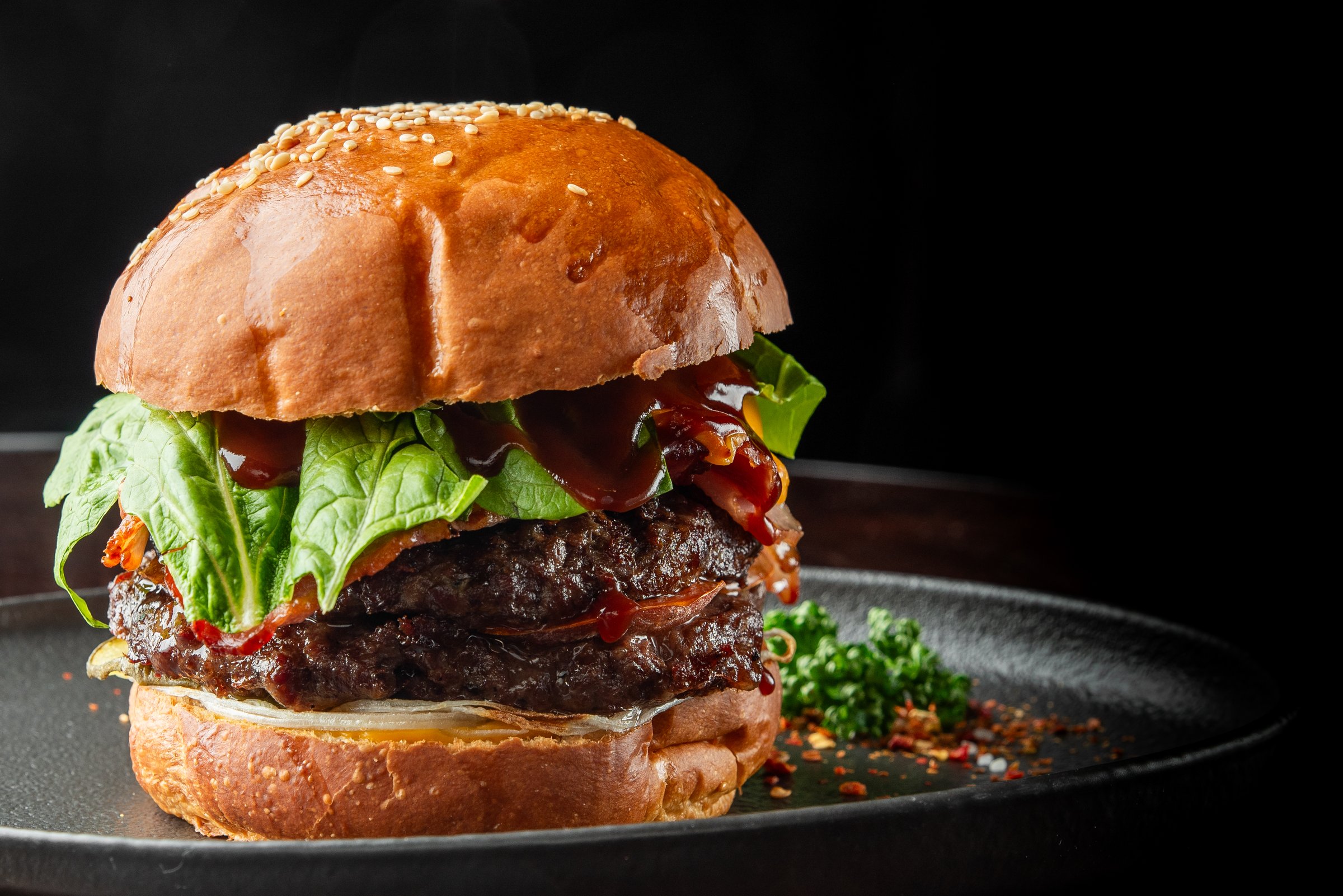 Gourmet burger with sesame seed bun, juicy beef patty, fresh lettuce, caramelized onions, and barbecue sauce on a black plate with parsley garnish. A delicious, high-quality food photography close-up.