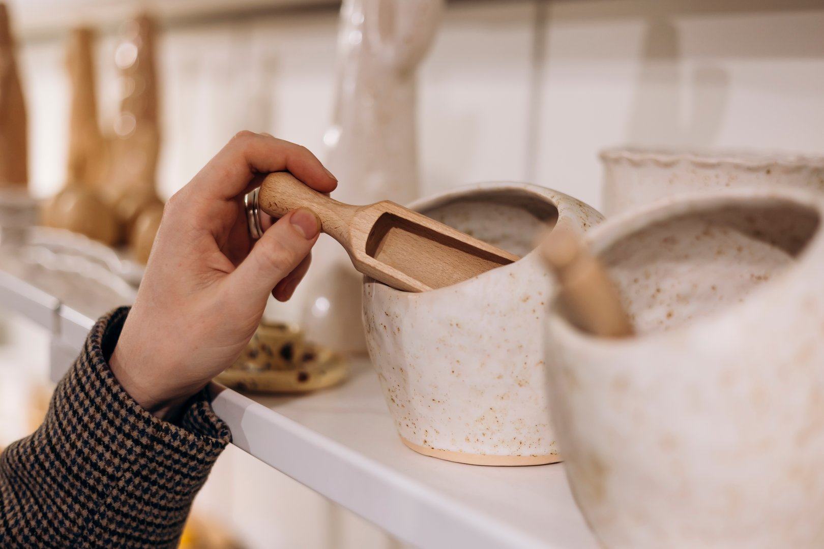 Close-up of a hand selecting a wooden spoon from a ceramic storage jar on a kitchenware shelf, highlighting mindful shopping and product choice in a clean retail setting.