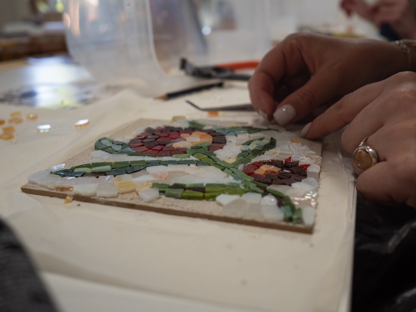 Her delicate hands, adorned with pastel nail polish and a wedding ring, move with precision and care. The flower pattern of the mosaic, which she is reproducing from an ancient design, highlights the feminine touch and attention to detail in her work. The scene has a sense of creativity and craftsmanship, emphasizing the personal and elegant nature of replicating historical artistry.