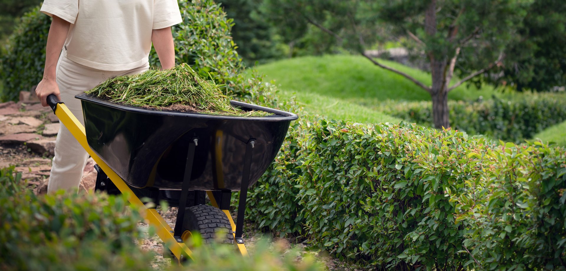 A gardener using a wheelbarrow filled with greenery in a beautifully maintained garden with lush surroundings.