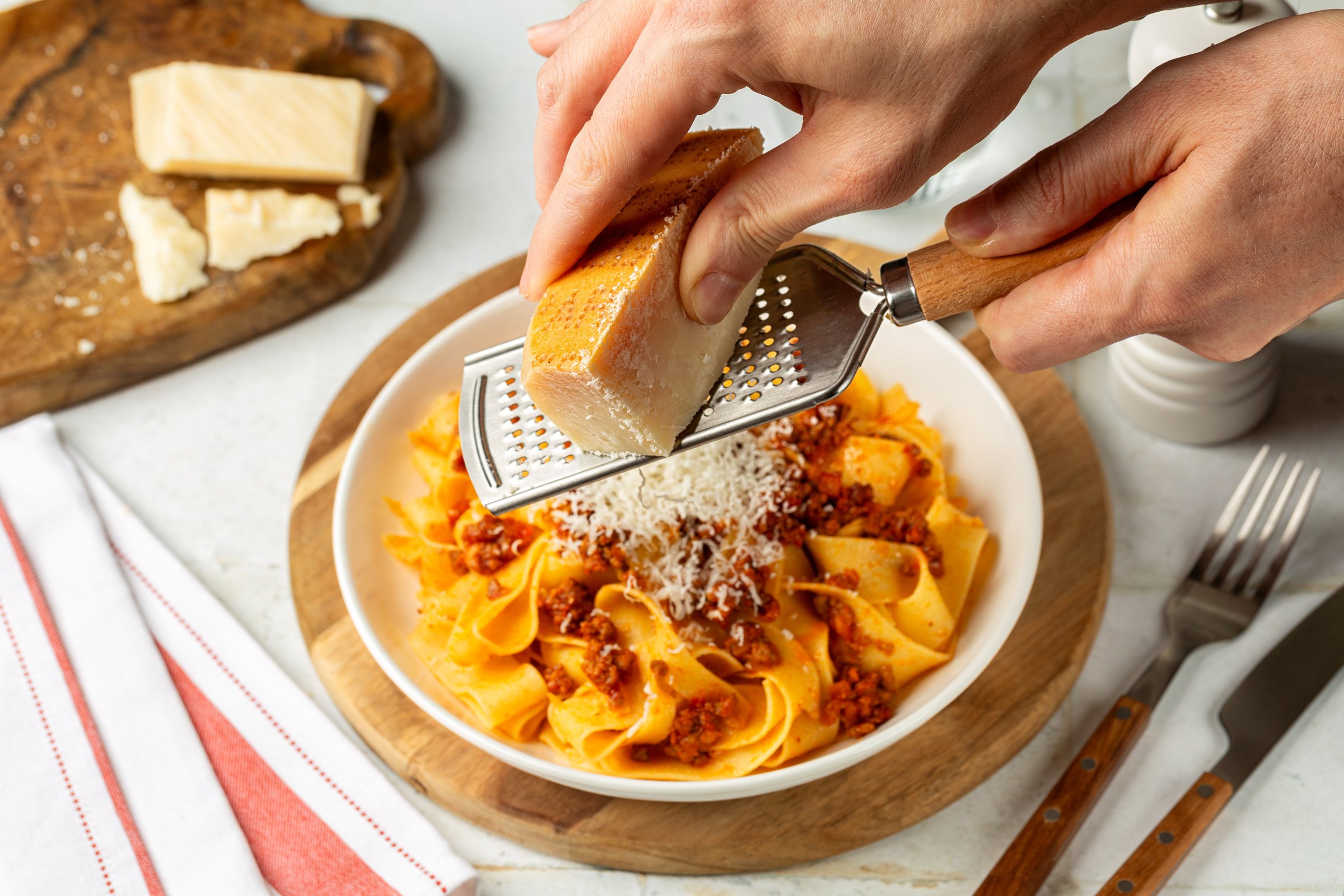 Man hands grating hard parmesan cheese on a pappardelle bolognese, or italian egg pasta with meat ragu sauce. White table. Selictive focus on a grater and cheese.
