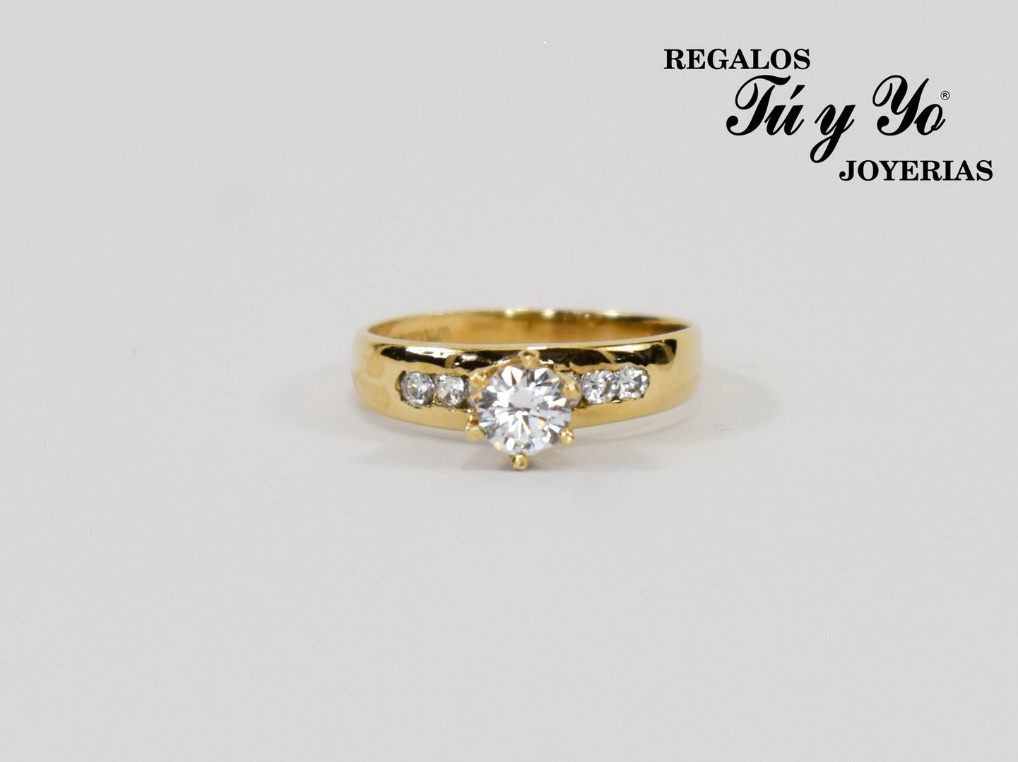 Gold ring with central diamond and smaller side stones, displayed against a white background. "Regalos Tú y Yo Joyerías" text.