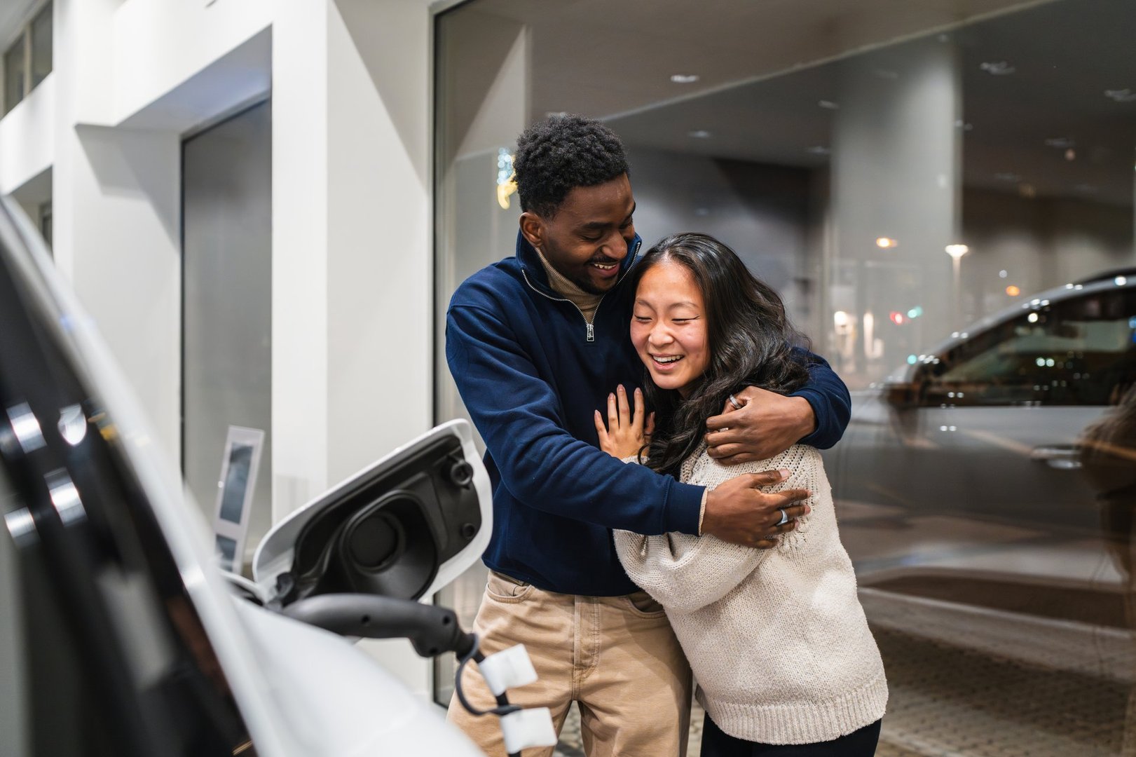 Young multi-ethnic couple hugging and celebrating their electric vehicle purchase at a car dealership, embracing eco-conscious choices