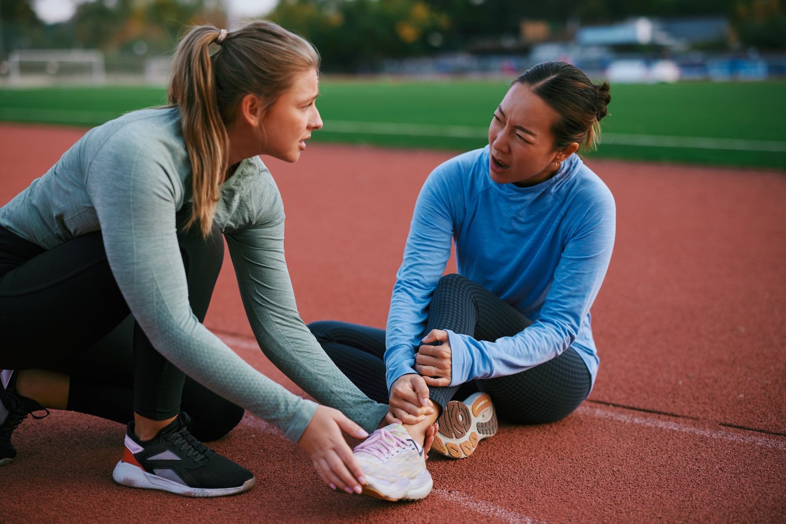 Two women in athletic wear sit on a running track, helping each other stretch an ankle before a workout. Outdoor fitness, teamwork, and motivation shine in this supportive moment.