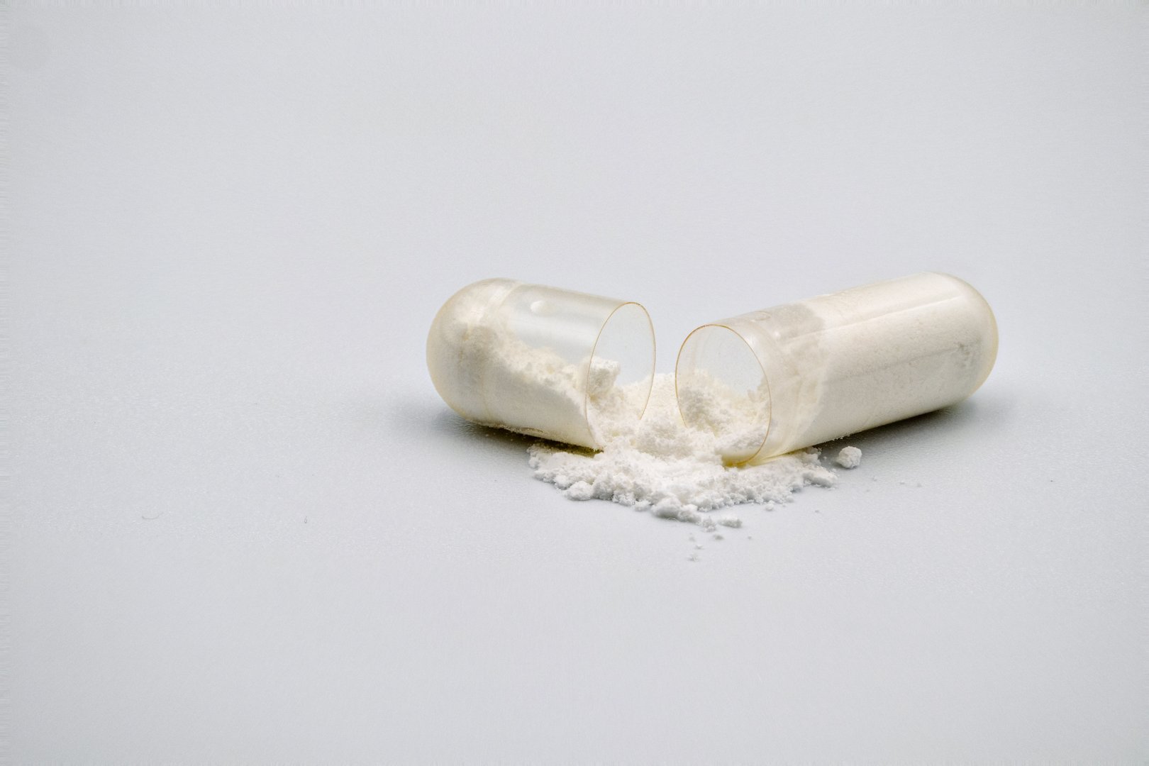 A clear, open capsule with white powder spilling out, resting on a pristine white surface, highlighting pharmaceutical contents.