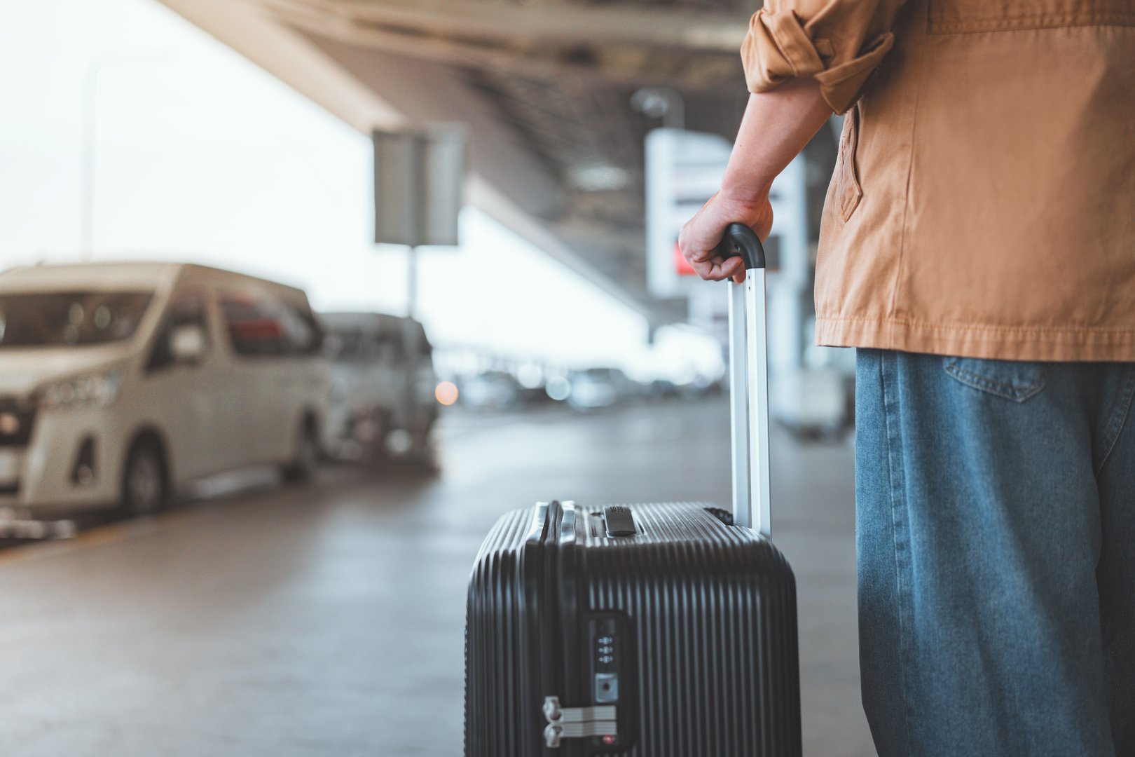 Rear view of male traveler standing with suitcase waiting for taxi at outside airport – travel concept, transportation and beginning of journey, copy space