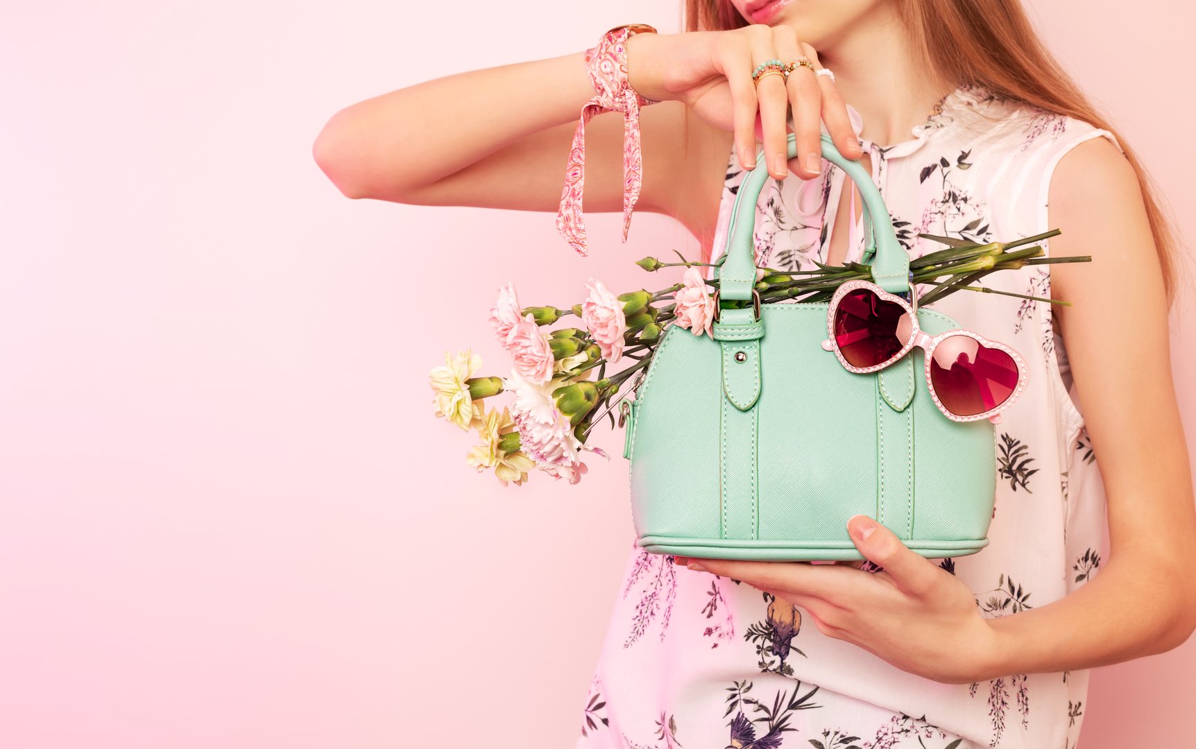Fashion spring accessories for woman. Girl holding small mint green handbag (purse), heart shaped sunglasses and flowers on pastel pink background. Free copy (text) space.