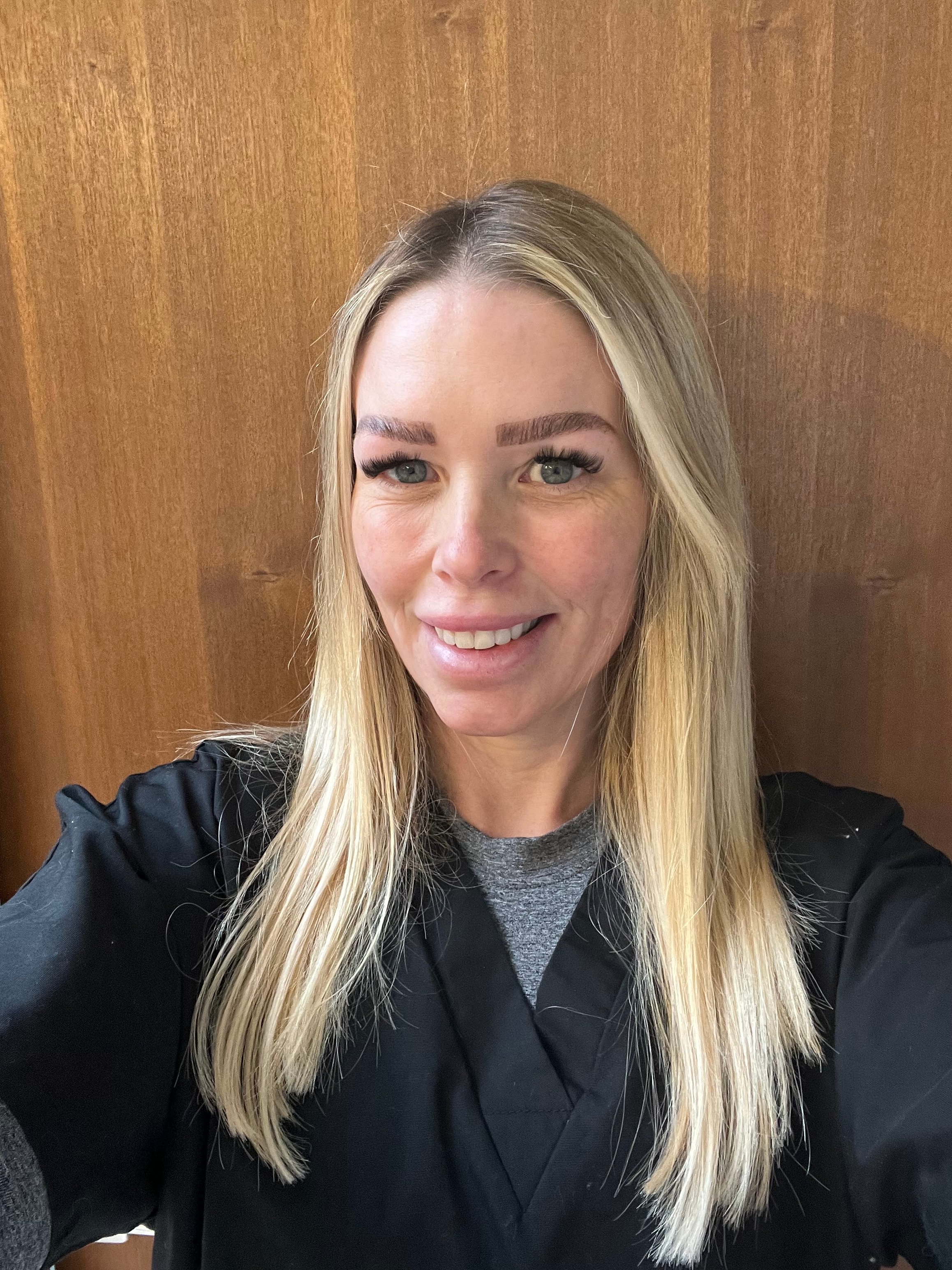 Person with long blonde hair wearing a black top, smiling in front of a wooden background.