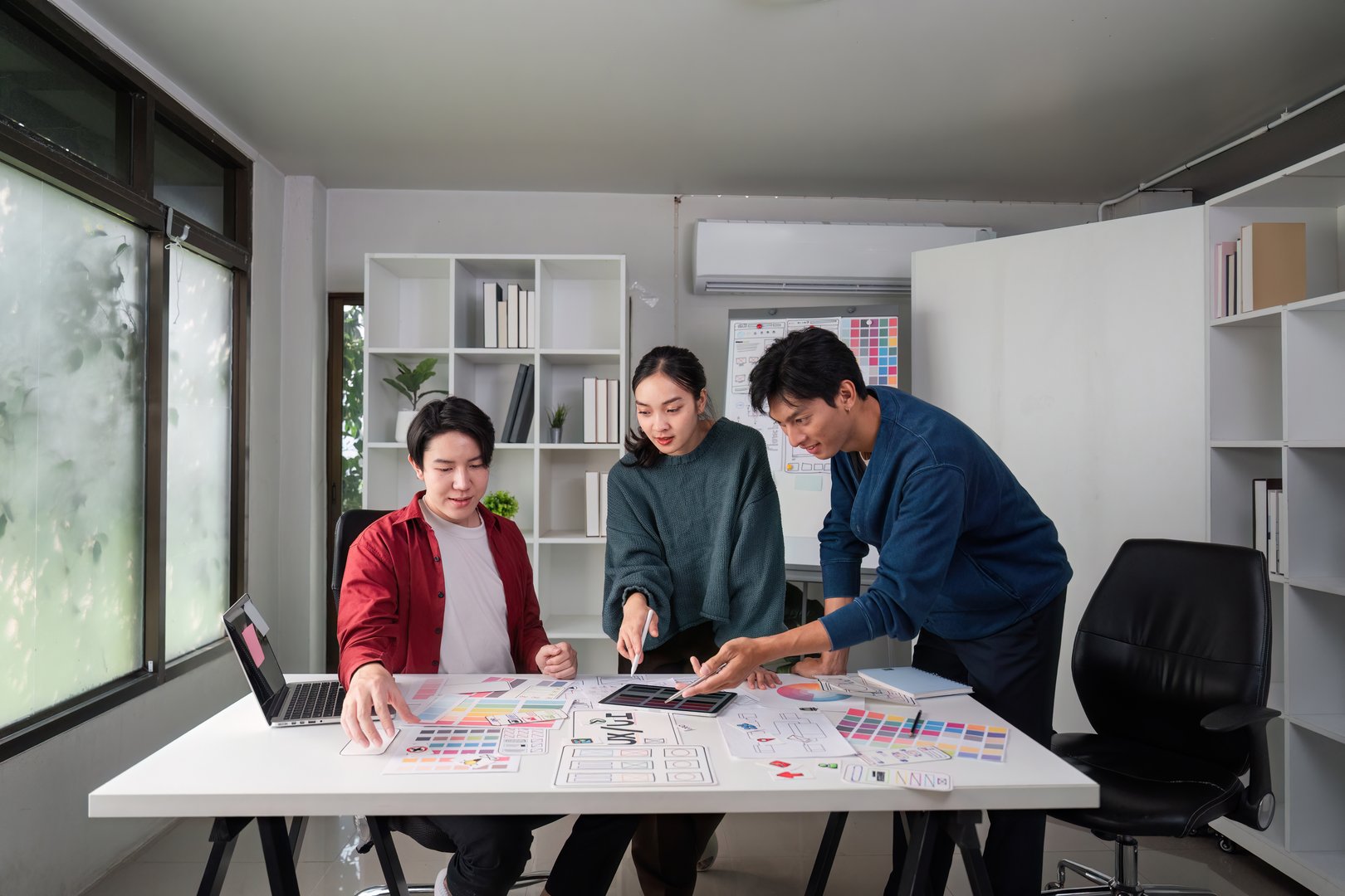 A team of three designers is actively collaborating on a UI/UX project, exchanging ideas and digital sketches in a bright workspace.