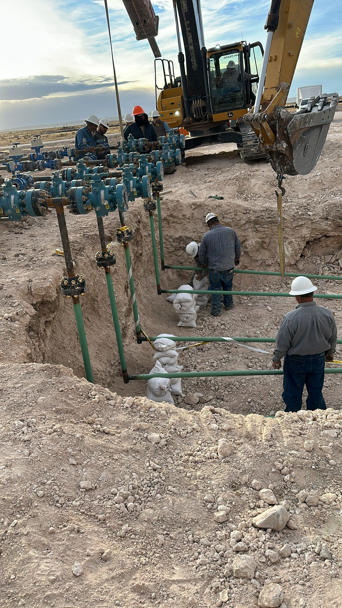 Workers in hard hats inspect underground pipes at a construction site with heavy machinery in the background.