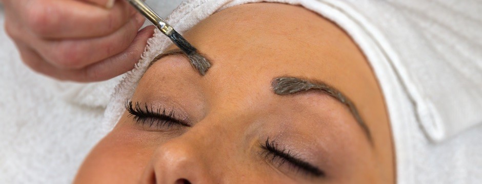 Close-up of a person getting eyebrow tint applied with a brush, eyes closed, wearing a towel wrap on their head.