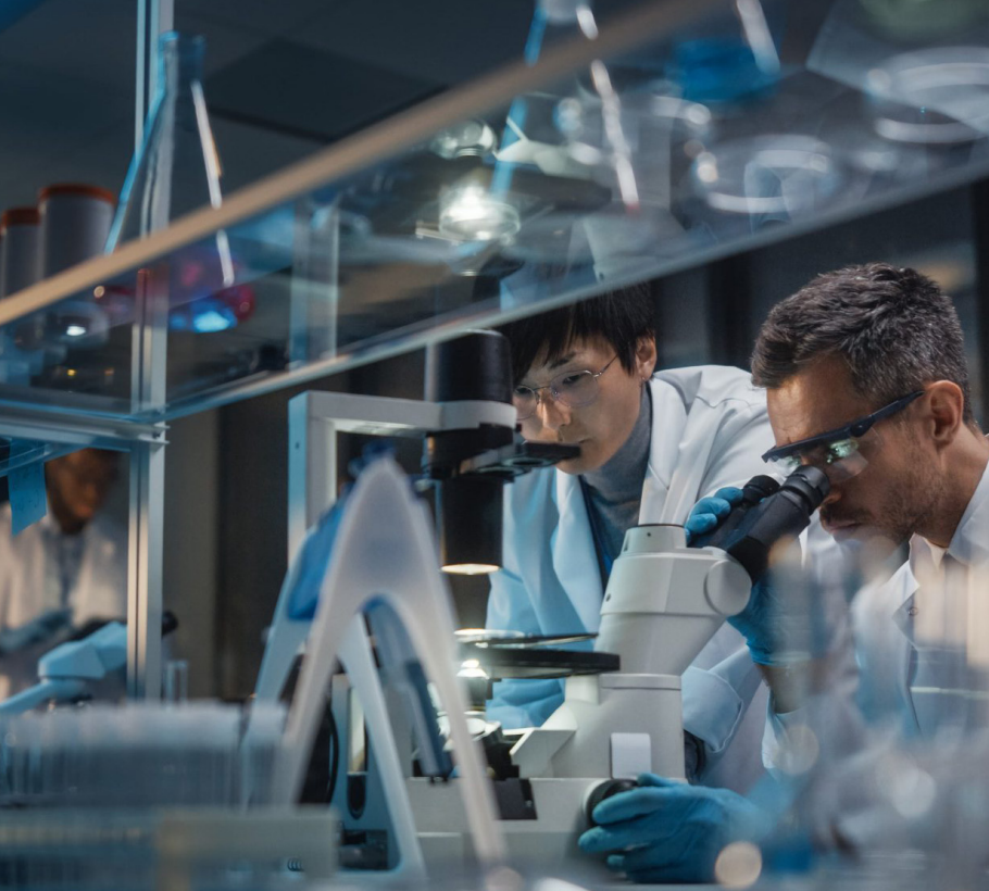 Scientists in a lab wearing white coats and gloves, using microscopes and surrounded by glassware and equipment.