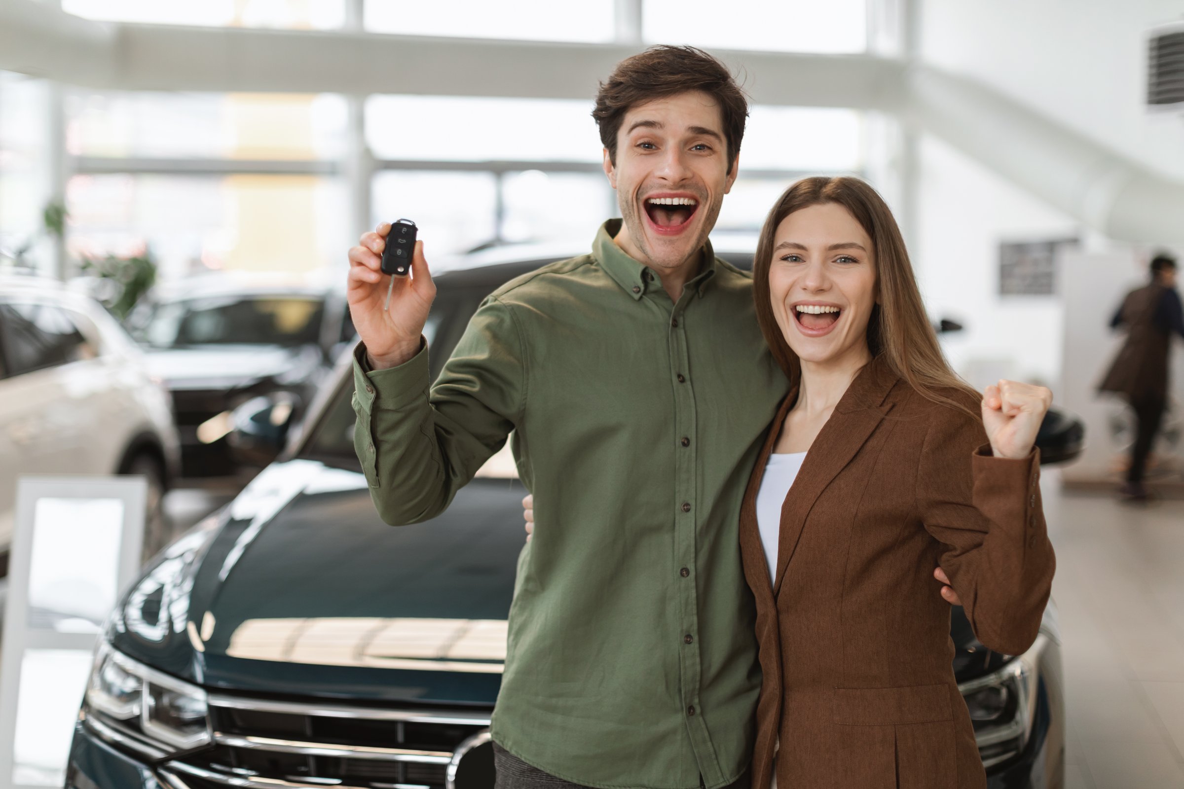 Overjoyed young Caucasian couple making YES gesture, showing car key, buying new auto at dealership, copy space. Excited millennial family celebrating purchase of luxury vehicle at showroom