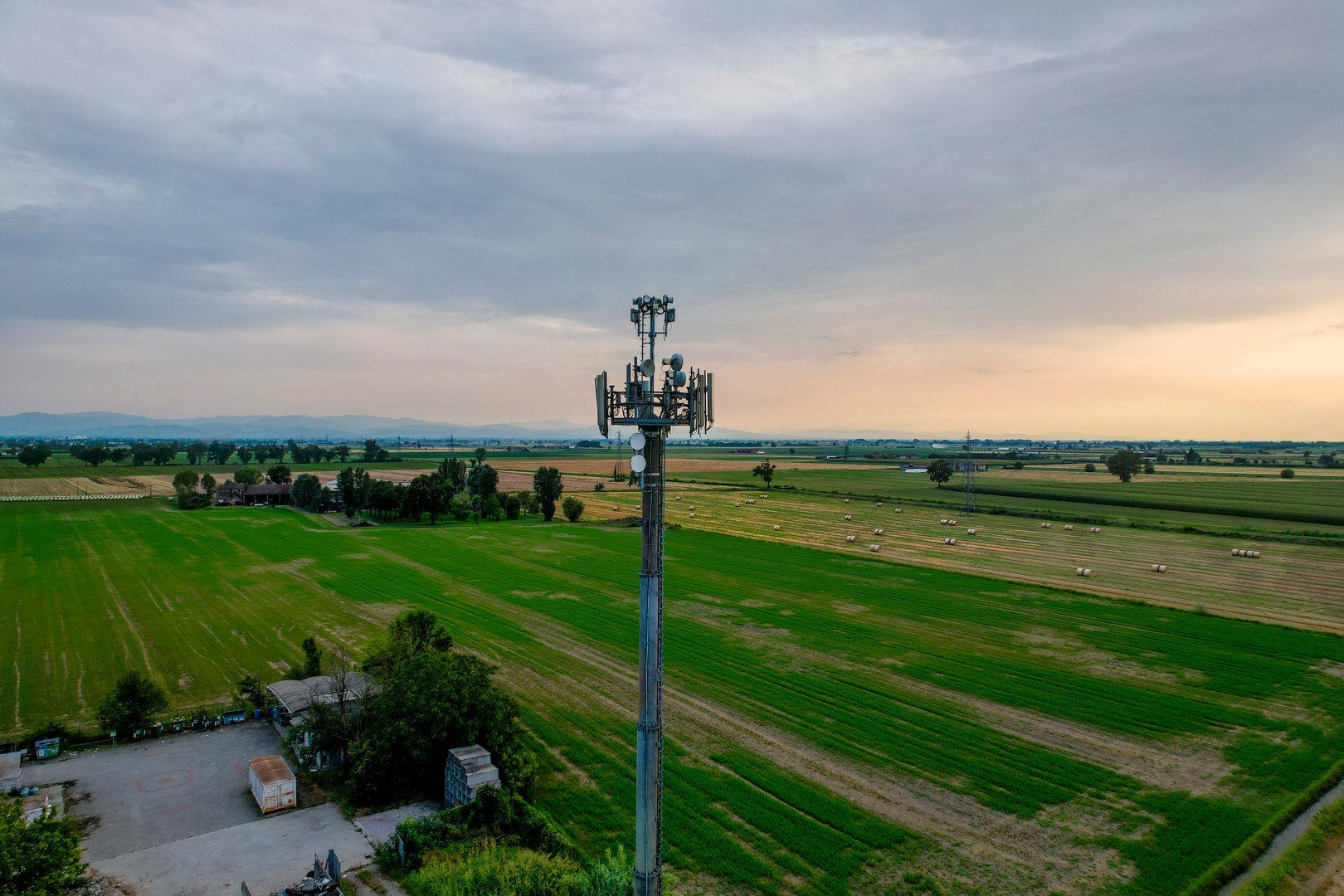 Aerial, A telecommunication tower is standing in cultivated green fields at sunset, providing signal coverage to a rural area. Drone view, pov