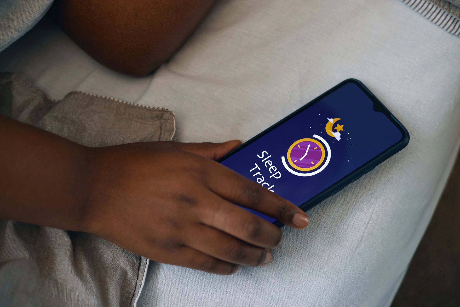 Hand resting on bed while holding smartphone with screen displaying sleep tracking app.