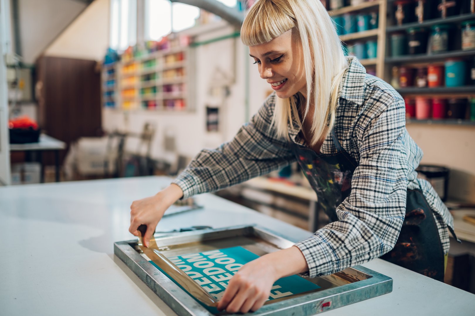 Smiling female graphic technology expert taping silkscreen printing plate with duck tape at printing workshop. Happy printing shop female worker preparing silkscreen printing press for printing.