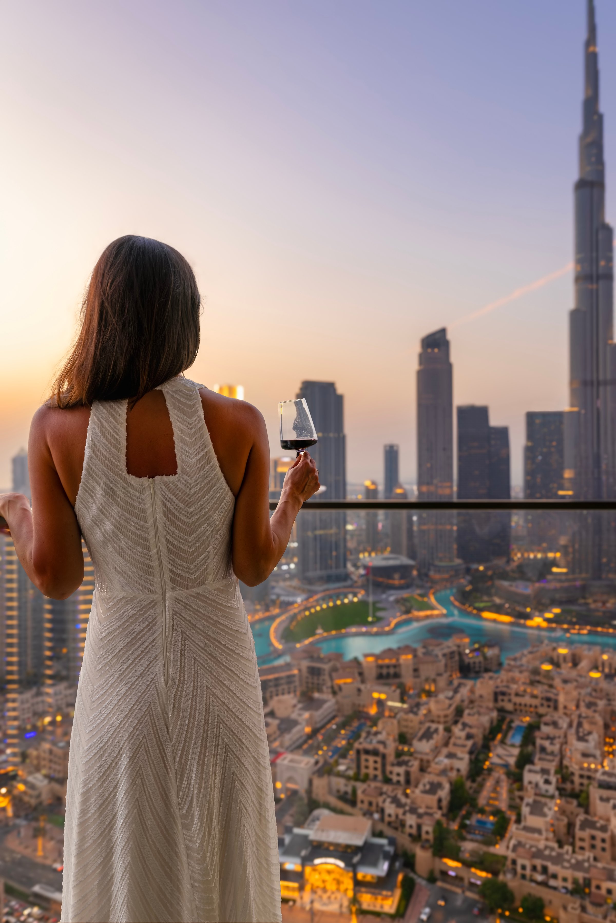 A beautiful woman in a dress enjoys the evening view of the illuminated Downtown Dubai skyline, UAE, with a drink in her hnd